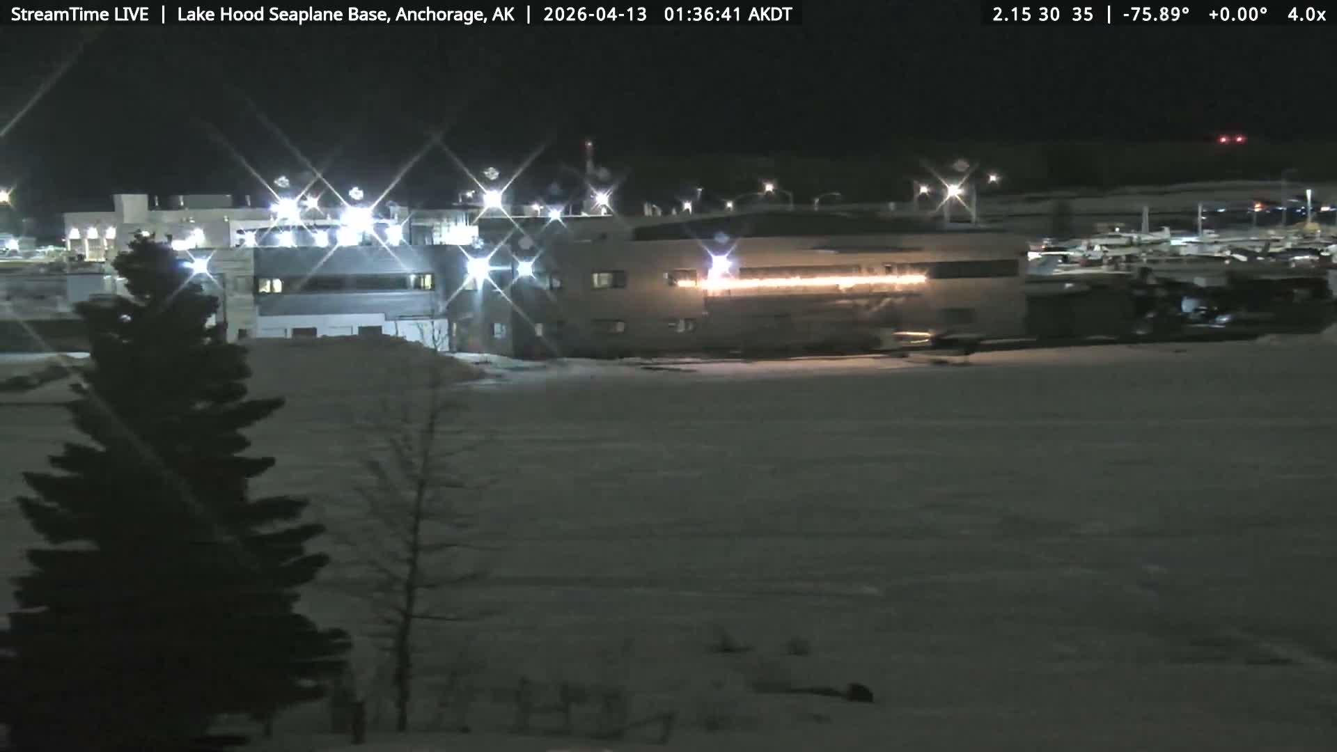 A clear night illuminates a striped building in the foreground, with numerous brightly lit structures and a parking area visible in the background at what appears to be an industrial or airfield complex.