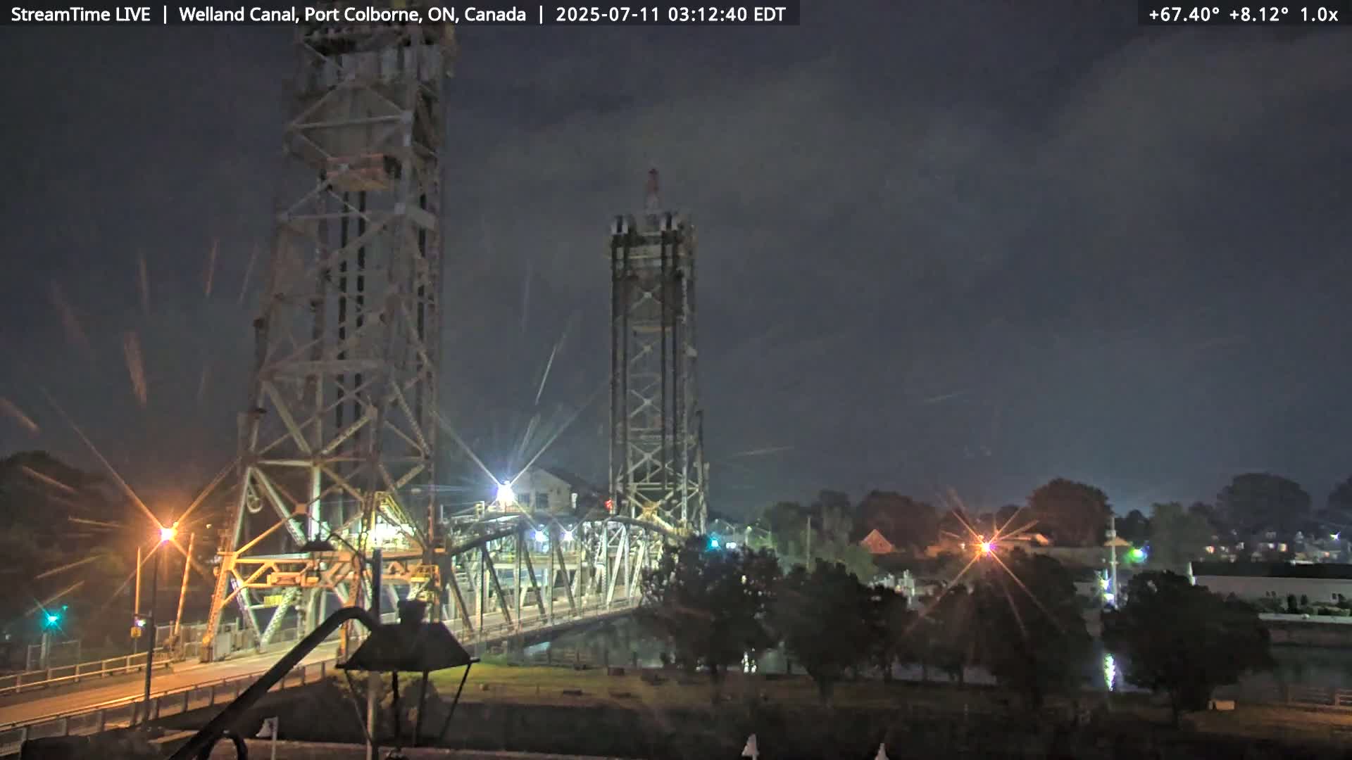 A nighttime view of a tall metal bridge over a dark canal under a slightly cloudy sky.