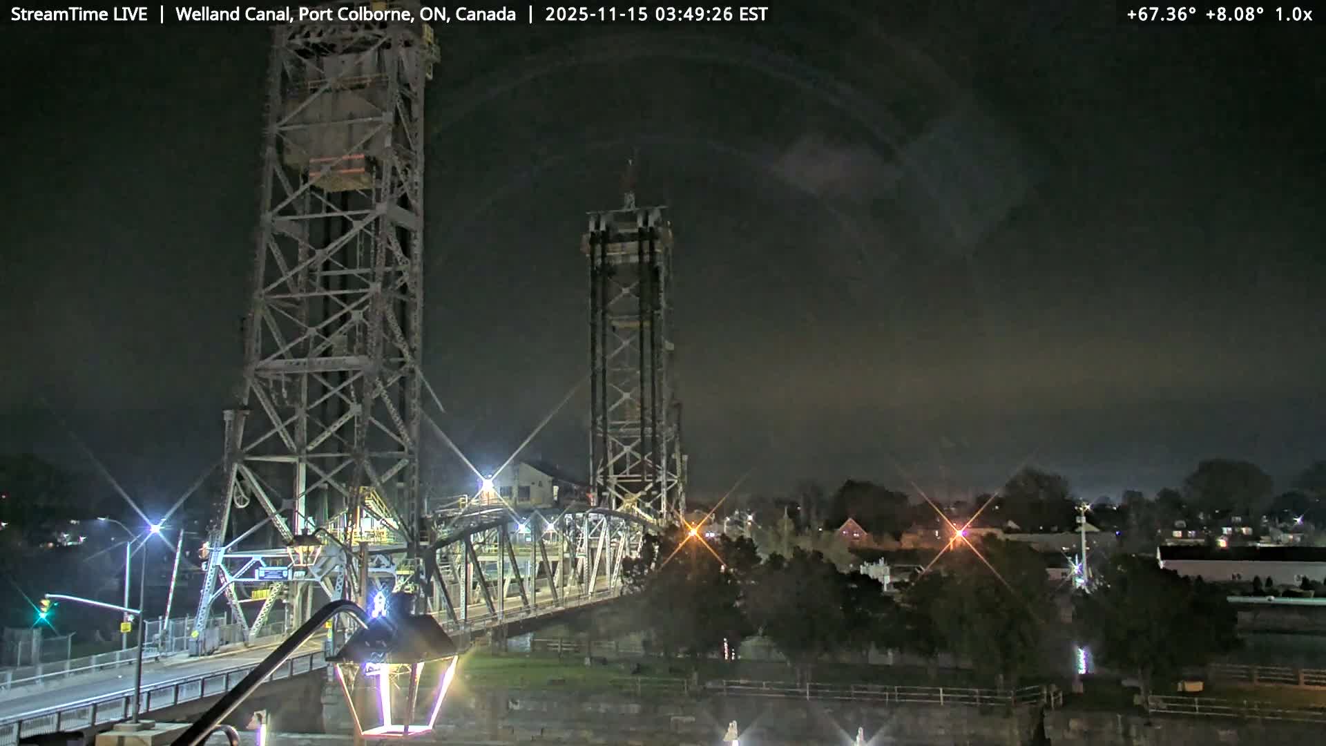 A large steel truss bridge with tall towers spans a dark waterway at night, surrounded by illuminated buildings and trees under an overcast sky.