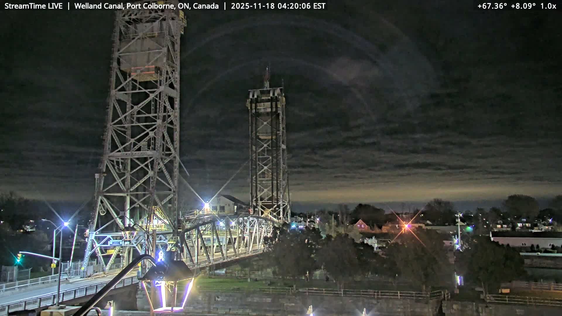 A large steel truss bridge with tall towers spans a dark waterway at night, surrounded by illuminated buildings and trees under an overcast sky.