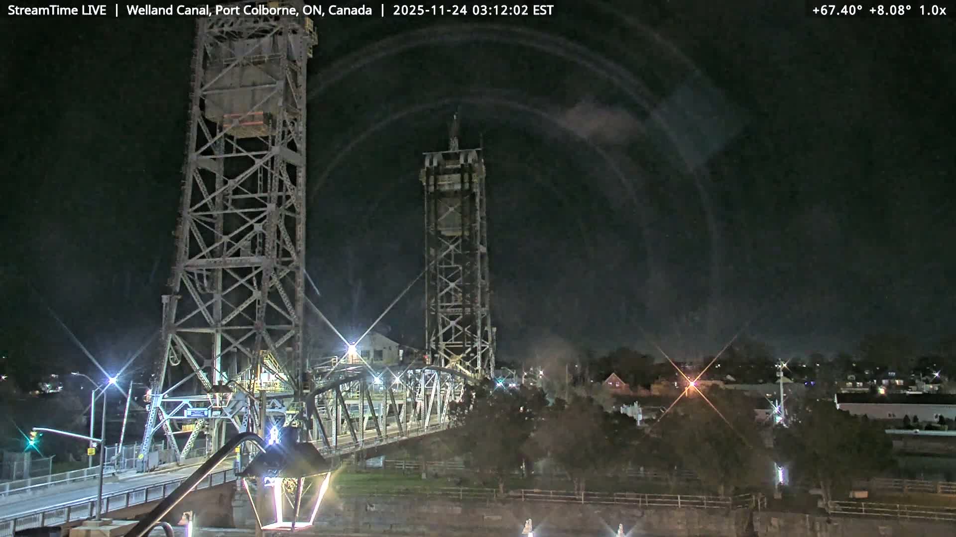 An illuminated steel truss lift bridge, with its tall towers and spanning roadway, crosses a dark waterway at night, casting light onto its structure and surrounding land, while distant town lights and a patch of mist are visible under a clear dark sky.