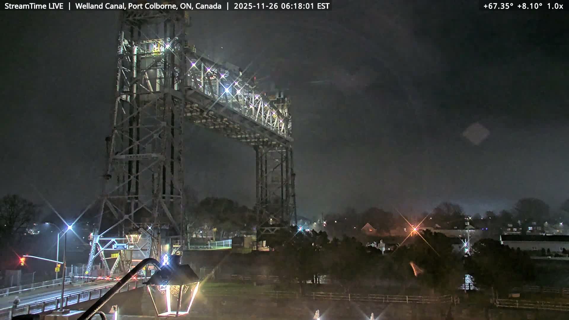 An illuminated steel truss lift bridge, with its tall towers and spanning roadway, crosses a dark waterway at night, casting light onto its structure and surrounding land, while distant town lights and a patch of mist are visible under a clear dark sky.