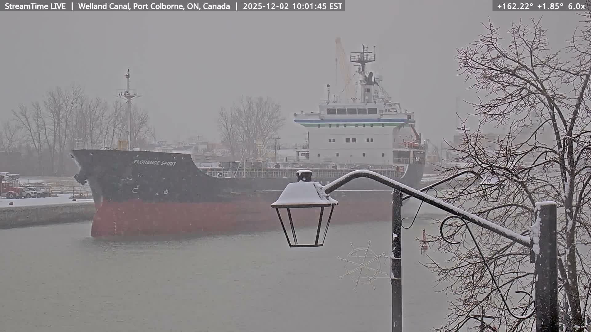 A cargo ship named "FLORENCE SPIRIT" navigates a grey canal during a snowfall, with snow accumulating on a lamppost and bare tree branches in the foreground.