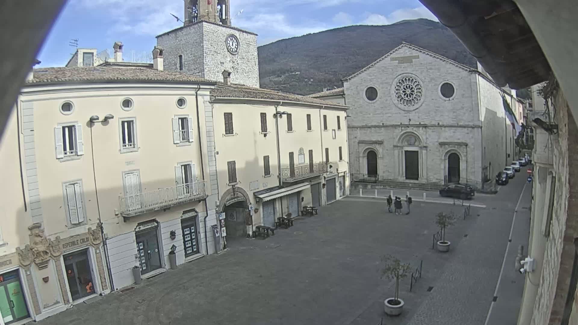 A mostly empty town square is seen on a partly sunny day, with light-colored buildings surrounding it, including a church with a rose window, and mountains in the background.