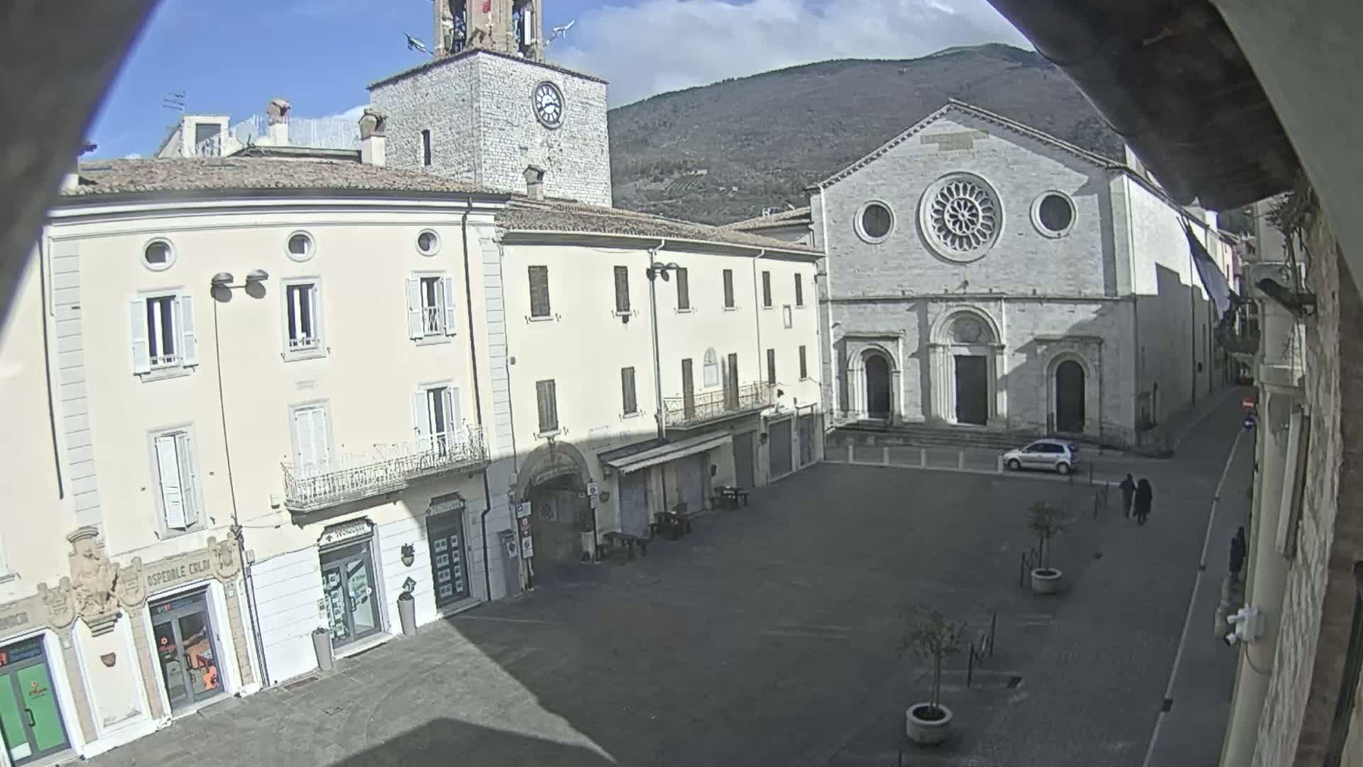 A sunny view of a town square, featuring a large church and several buildings, with mountains visible in the background.