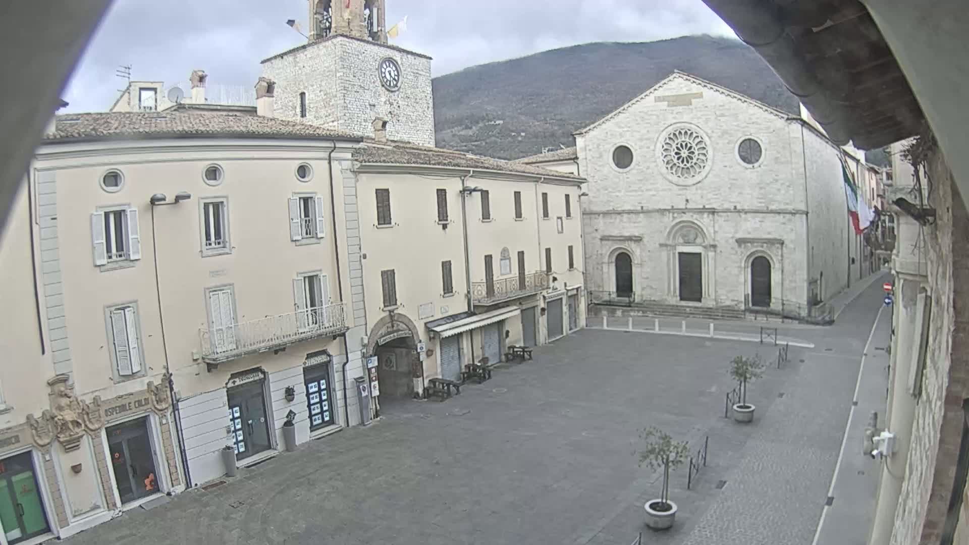 A mostly empty town square is shown on an overcast day, with several light-colored buildings surrounding it, including a church with a large rose window and a clock tower.