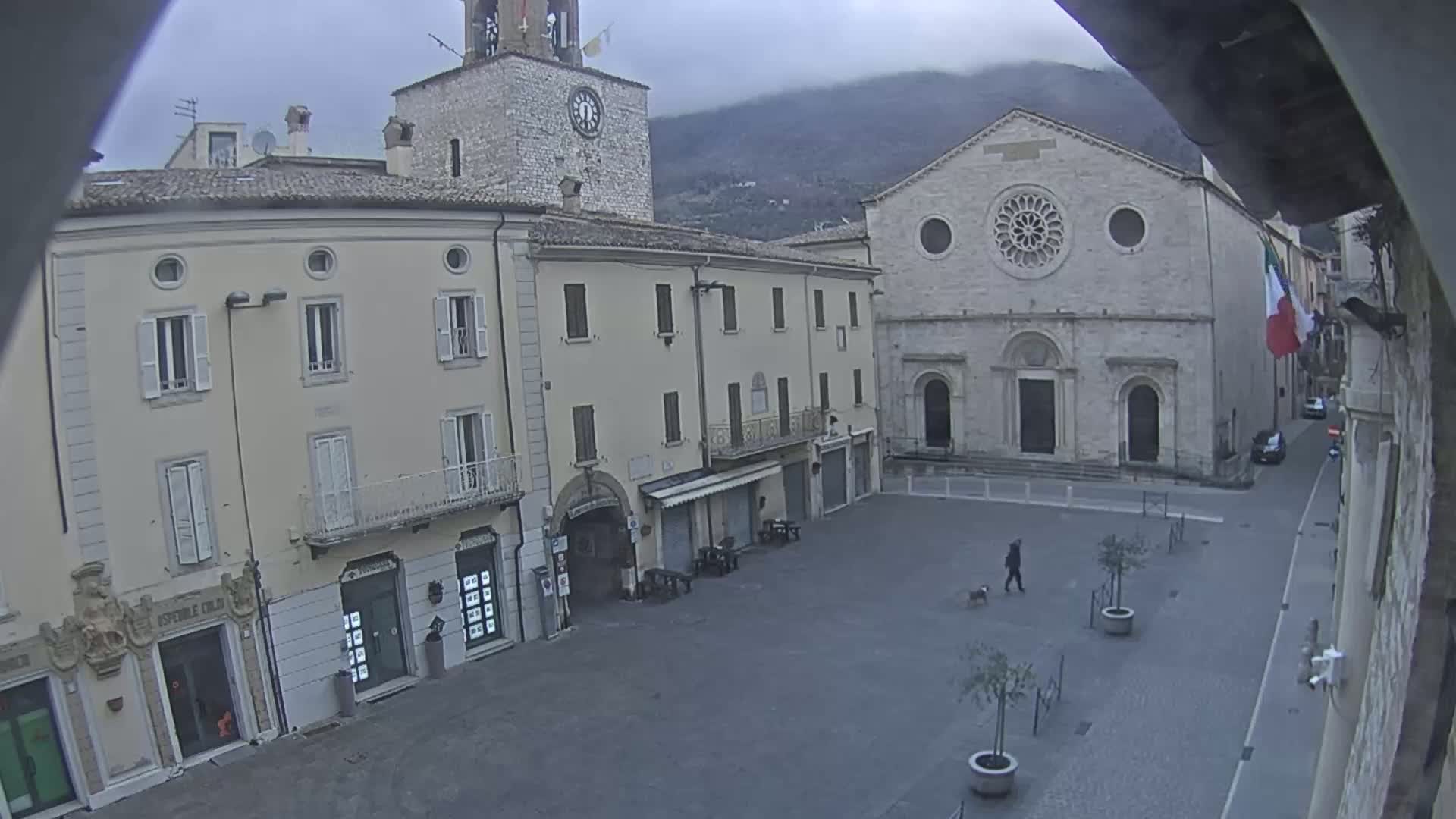 A mostly empty town square with stone buildings under an overcast sky, one building featuring a large rose window, and a person walking a dog.
