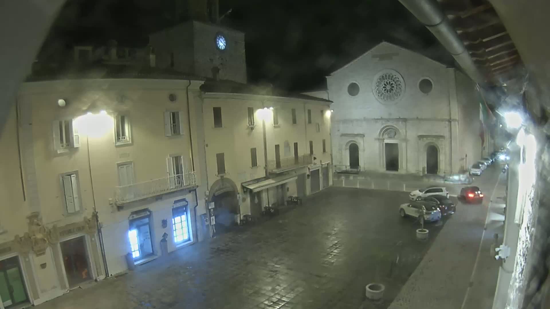 A nighttime view of a town square with several buildings, including a church with a rose window, under a slightly overcast sky.