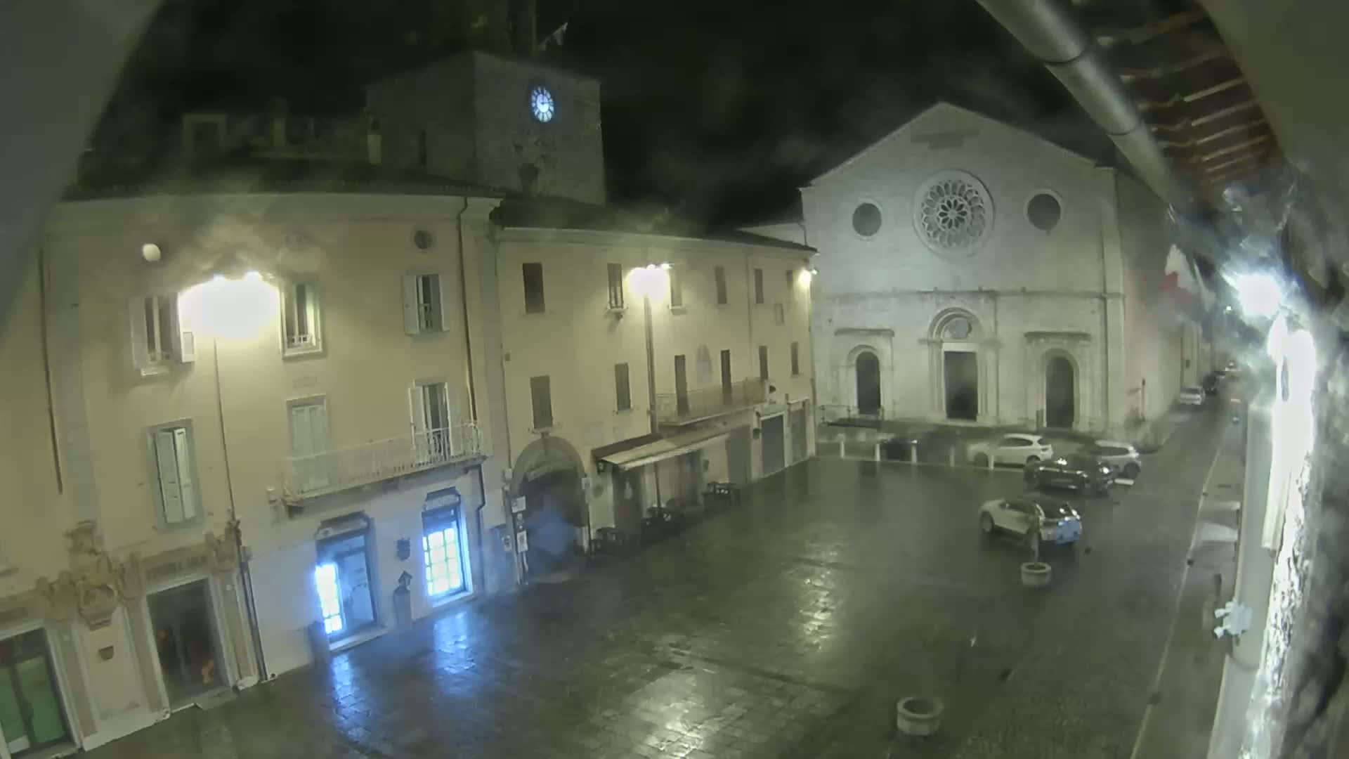 A rain-slicked town square at night is illuminated by streetlights, showing several buildings including a church with a rose window and a clock tower.