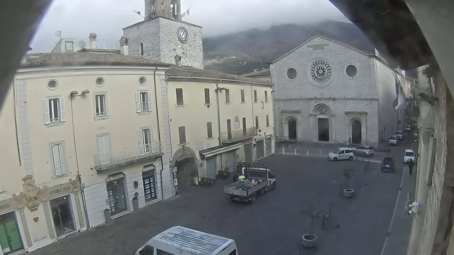 A town square, under an overcast sky, is surrounded by light-colored buildings, including a church with a rose window, and contains several parked cars and a couple of vans.