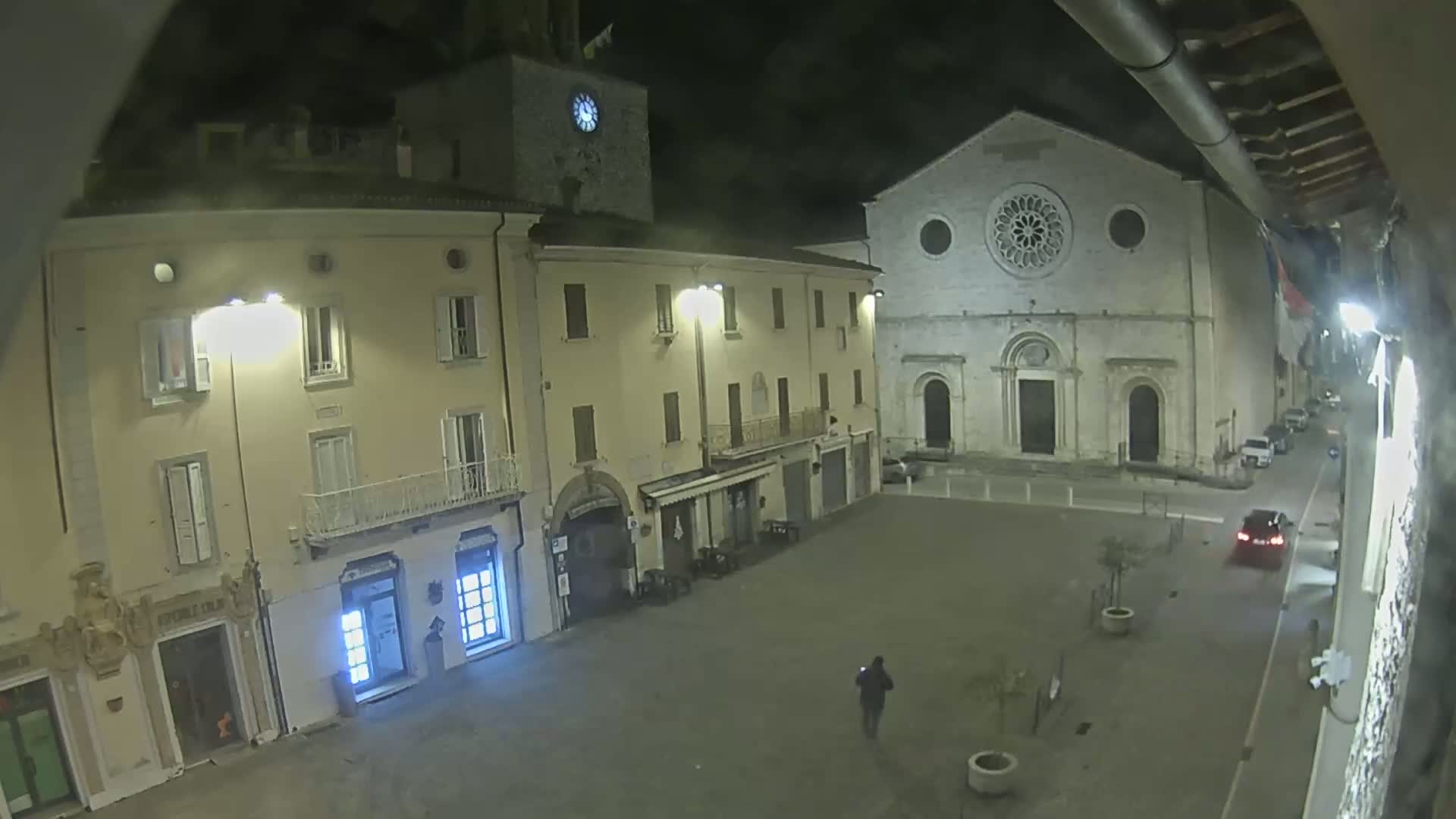 A nighttime view of a town square shows a large stone church, several buildings with illuminated windows, a person walking, and a car parked nearby under dark, clear skies.