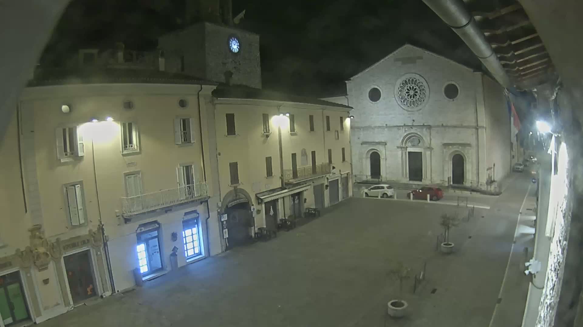A nighttime view of a town square with buildings, including a church with a rose window, under a dark, cloudy sky.