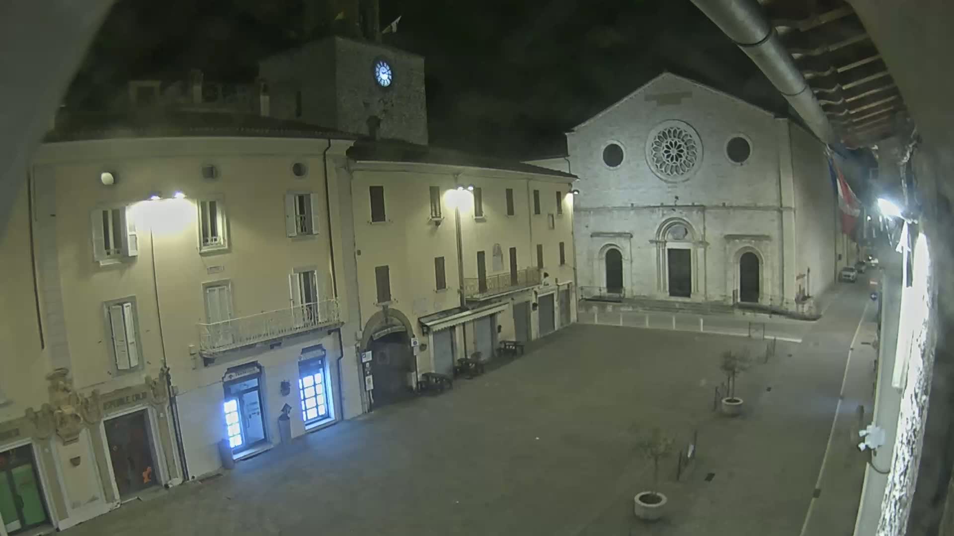 A nighttime view of a town square with buildings lit by streetlights, including a church with a rose window, and a clock tower.