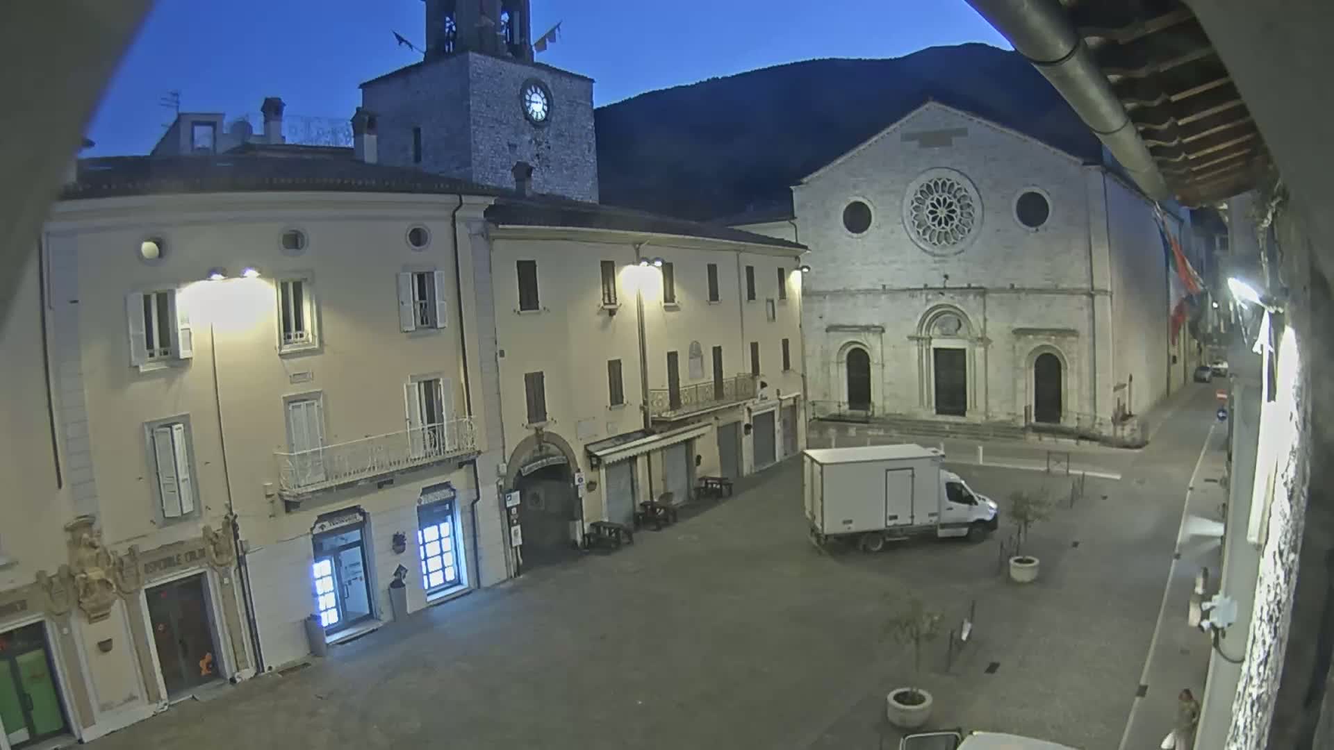 A nighttime, high-angle view of a town square, featuring illuminated buildings, including a church with a rose window, a clock tower, and a parked delivery van.