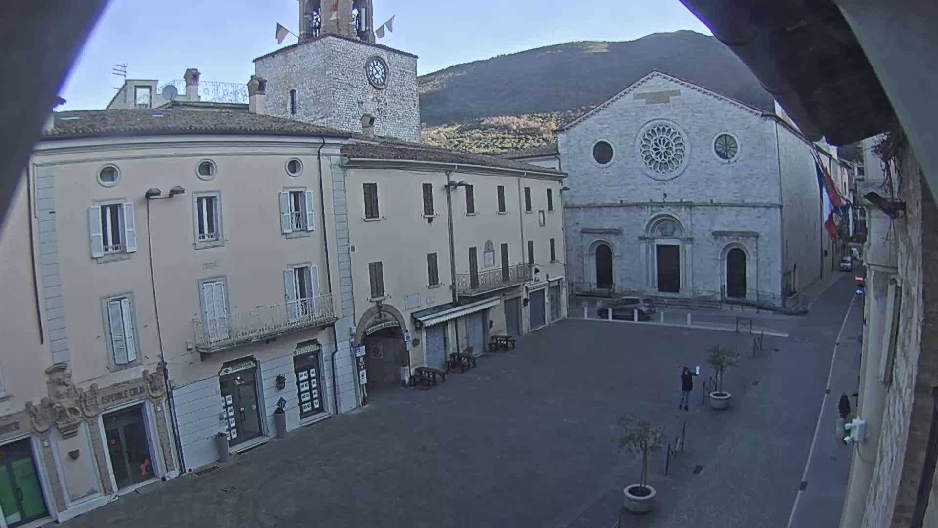 A mostly empty town square is seen under a clear, bright sky, surrounded by light-colored buildings, including a church with a large rose window and a clock tower.