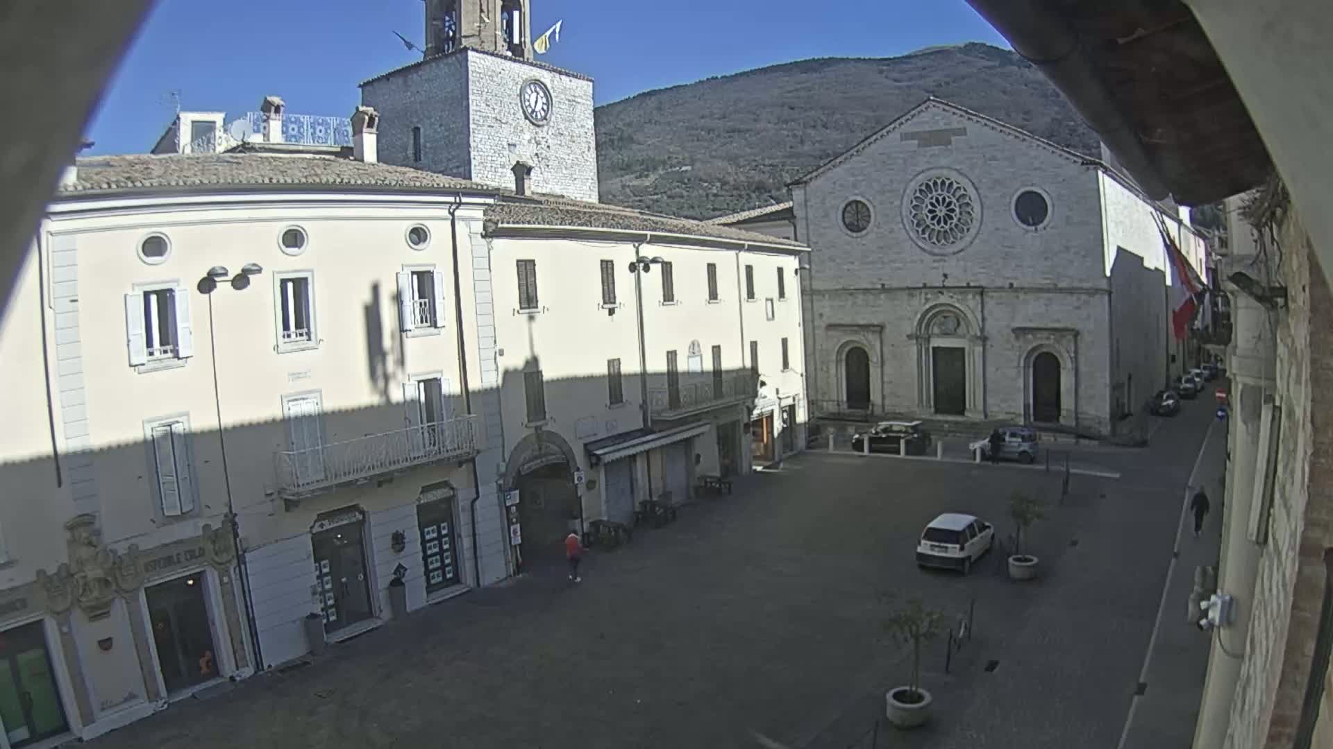 A mostly empty town square is seen on a sunny day, with several buildings surrounding it including a church with a rose window, a clock tower, and other shops and residences.