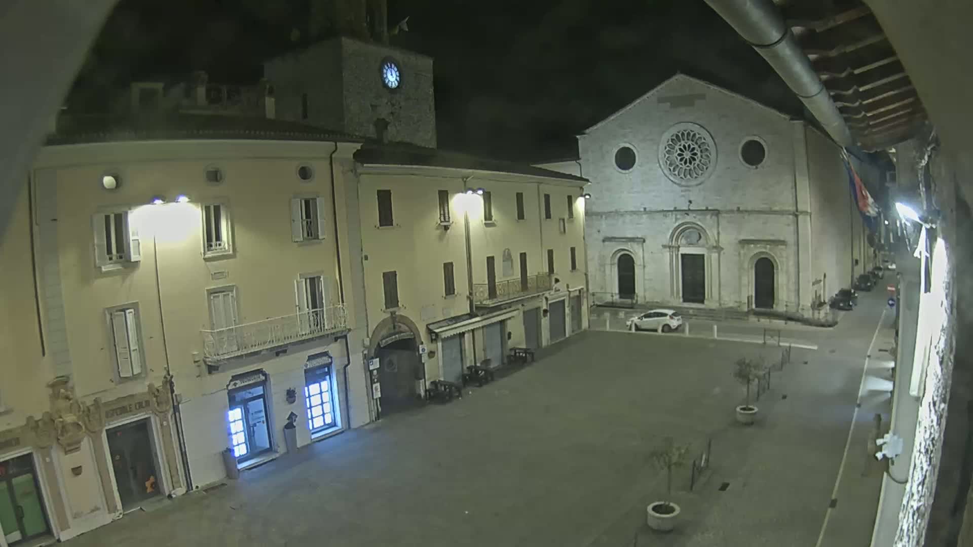 A nighttime view of an empty town square with buildings, including a church with a rose window, under dark skies.