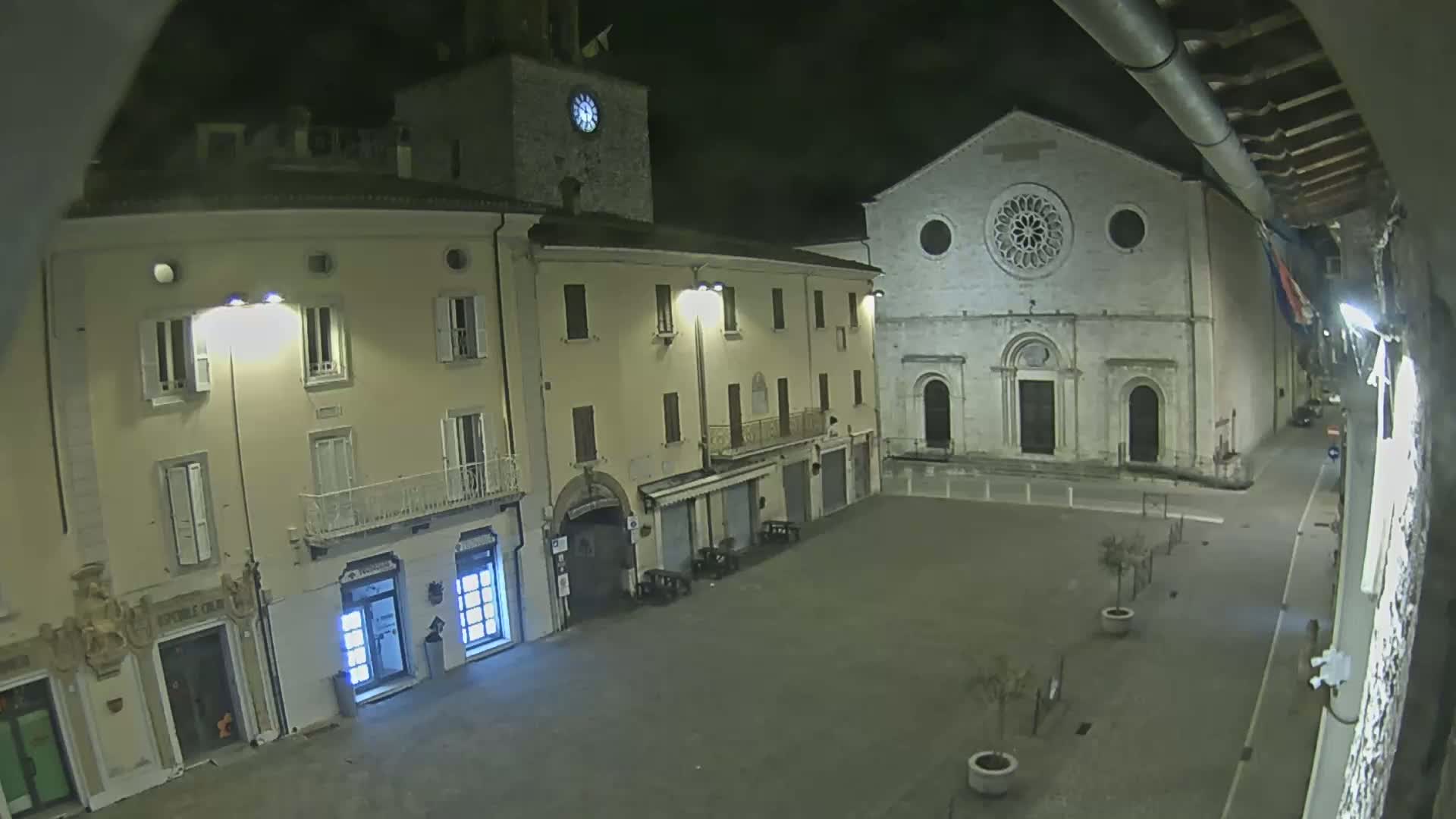 A nighttime, high-angle view of a mostly empty town square with buildings, including a church with a rose window, illuminated by streetlights.