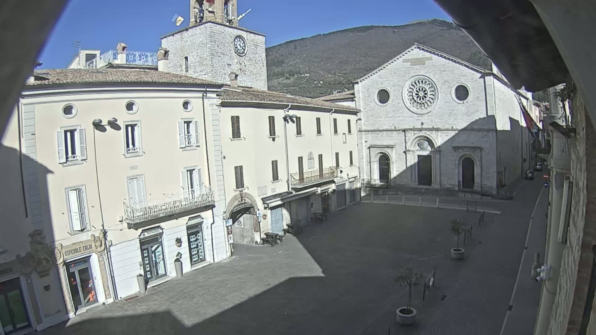 A sunny day reveals a town square with several buildings, including a clock tower and a church with a large rose window, backed by a mountain range.