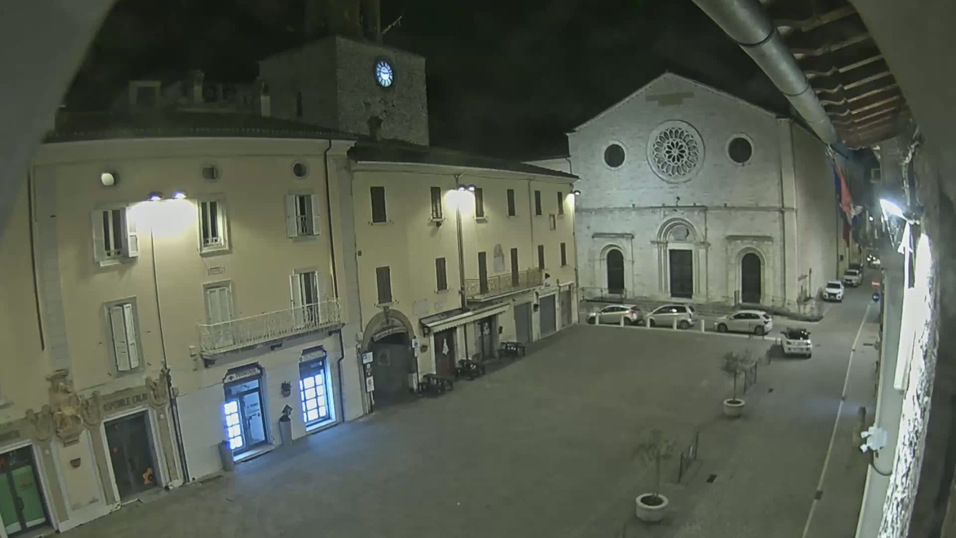 A nighttime view of a town square with several buildings, including a church with a rose window, under dark conditions.