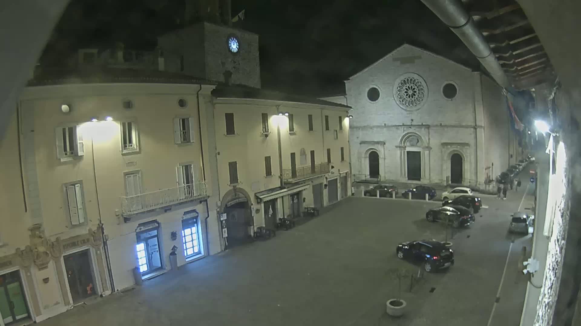 A nighttime view of a town square with several cars parked, buildings including a church with a rose window, and streetlights illuminating the scene.