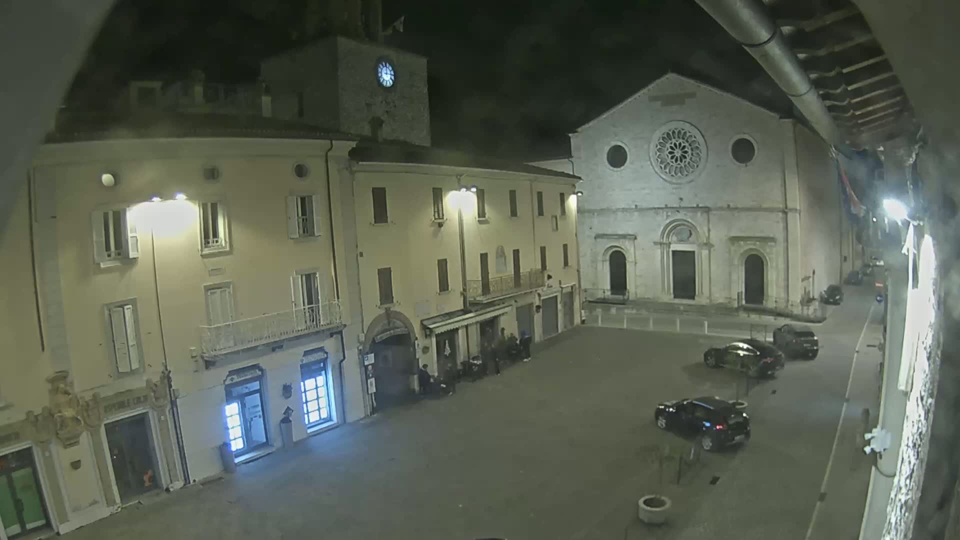 A nighttime view of a town square with several buildings, including a church with a rose window, and a few cars parked.