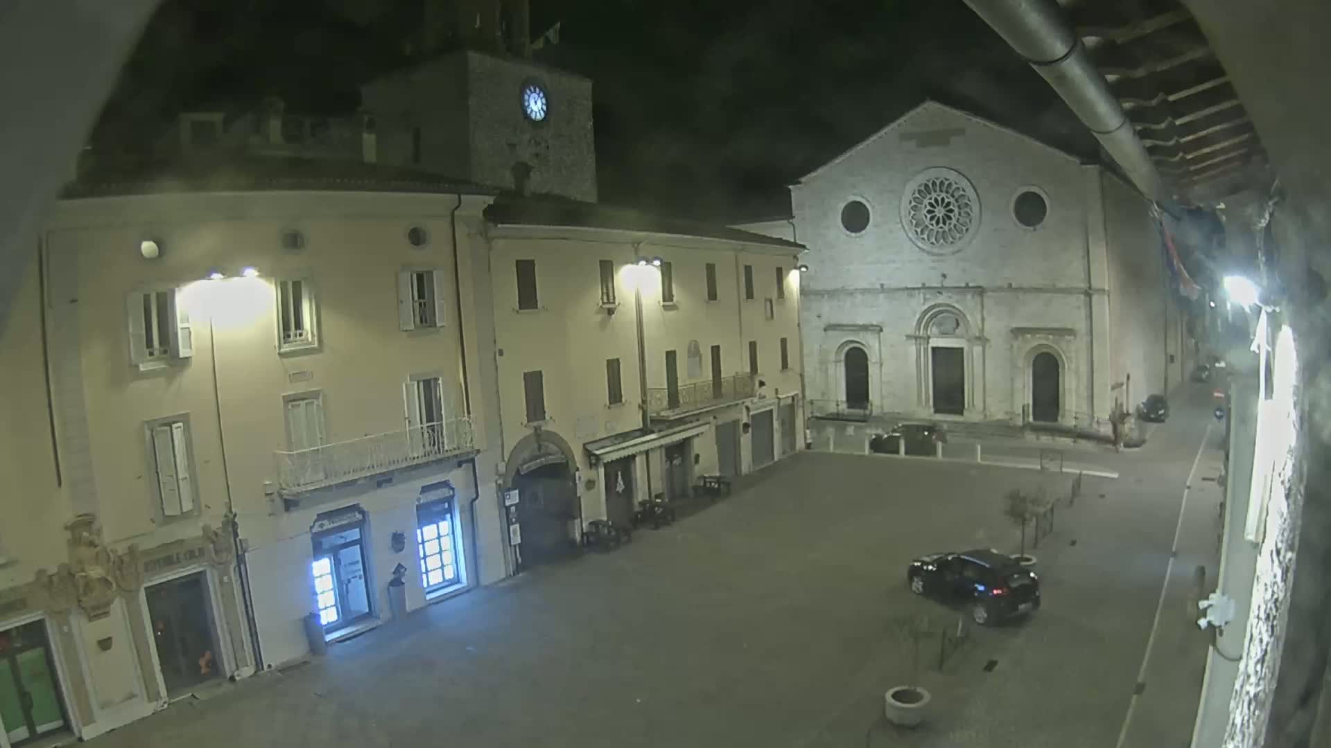A nighttime view of a town square with several buildings, including a church with a large rose window, under a dark, possibly cloudy sky.