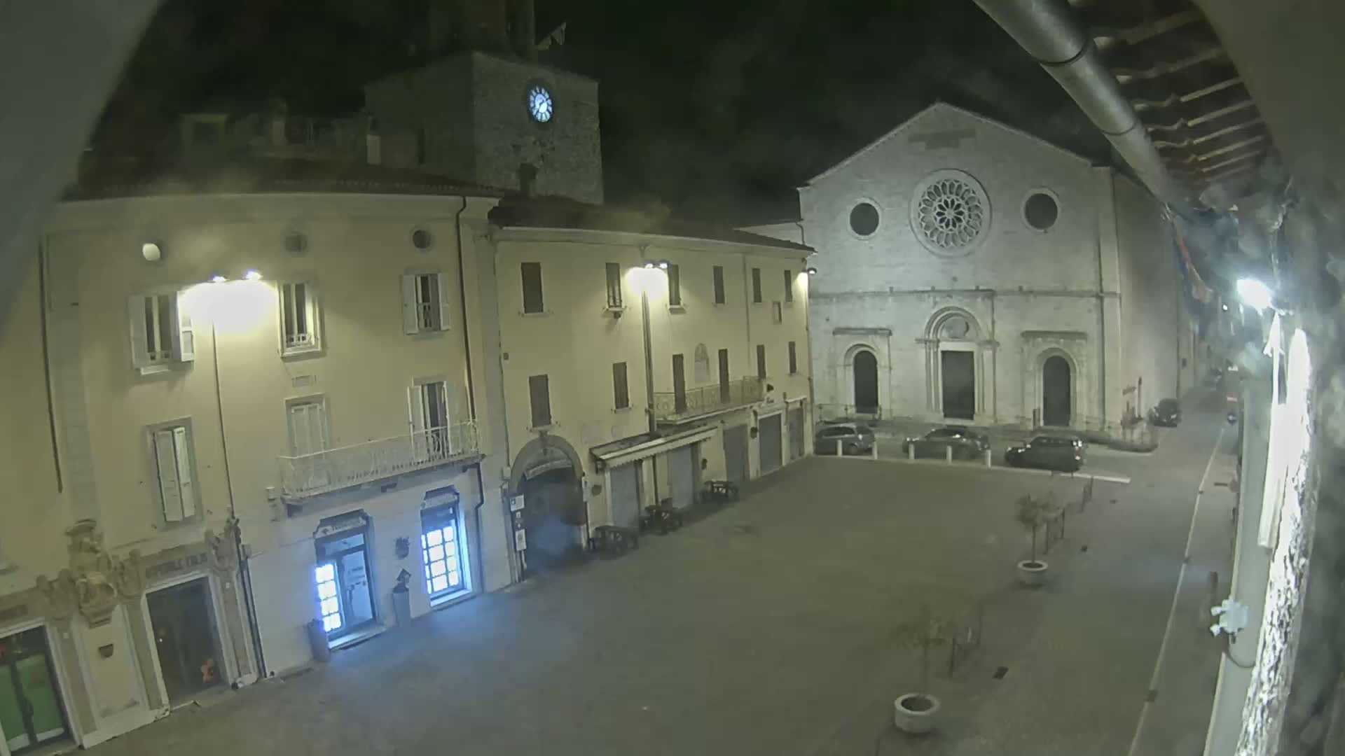 A nighttime view of a town square with several buildings, including a church with a rose window, under dark conditions.
