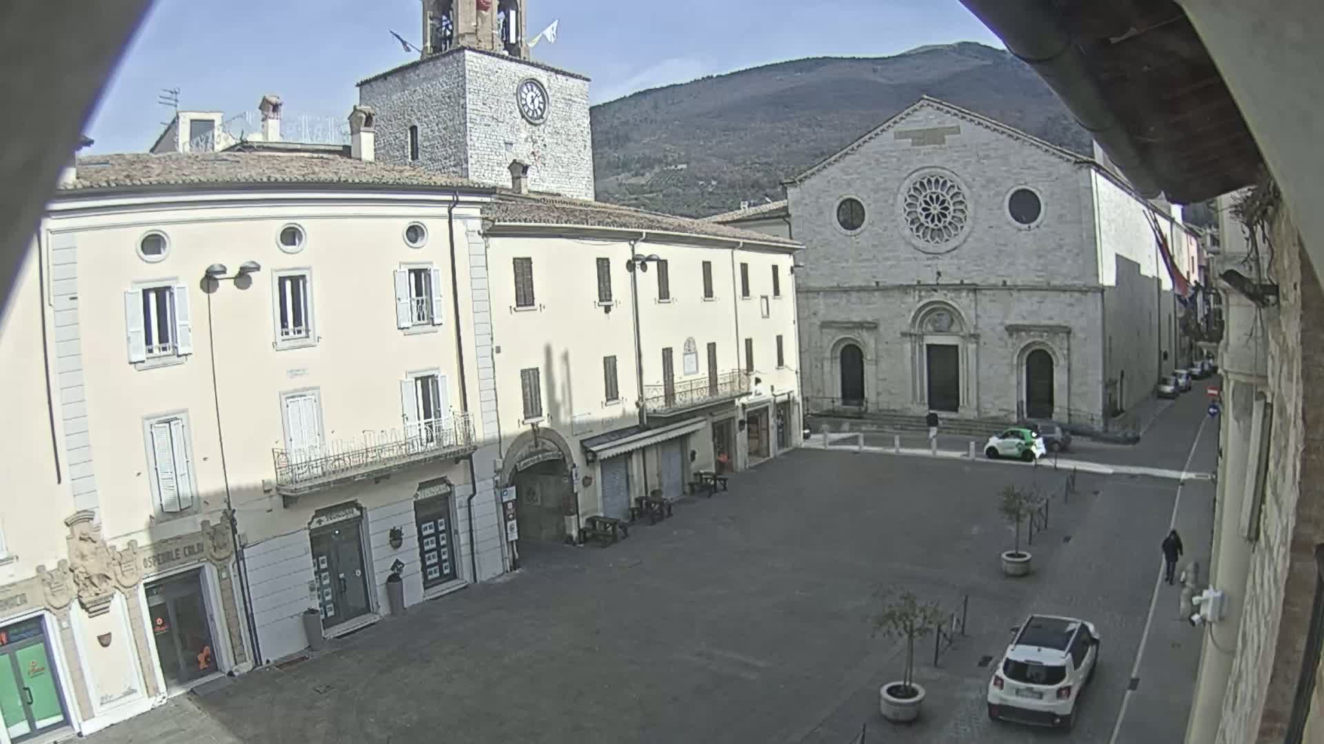 A mostly empty town square is seen on a sunny day, with a church and clock tower in the background, and a few cars parked in the square and nearby streets.