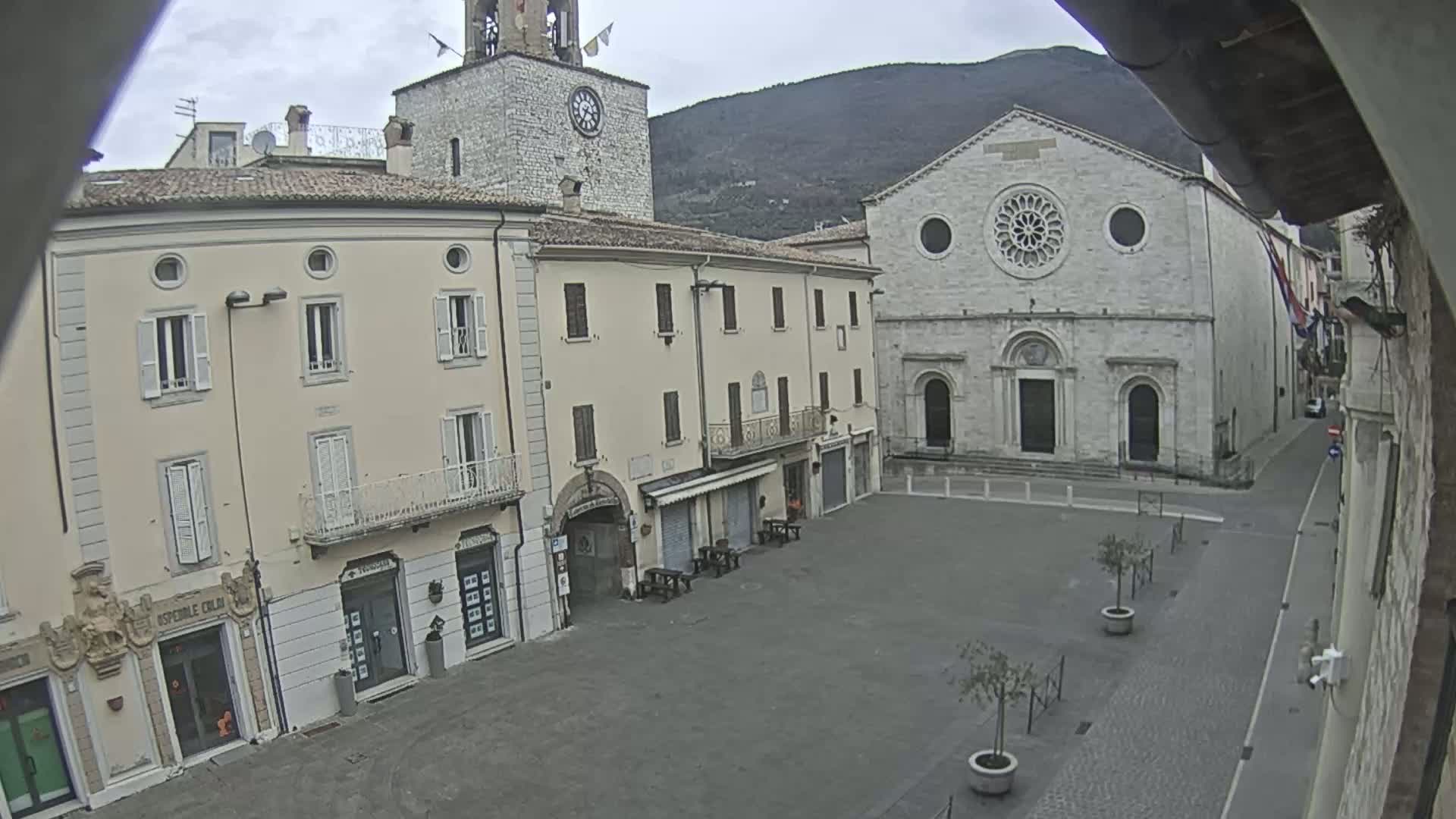 A mostly empty town square is seen on an overcast day, with several buildings surrounding it, including a church with a large rose window and a clock tower.