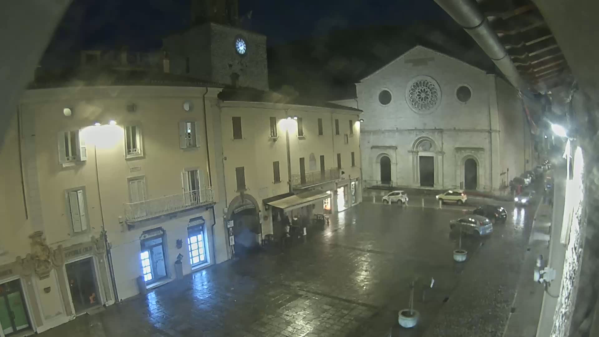 A rain-slicked town square at night, illuminated by streetlights, features several buildings including a large church with a rose window and a clock tower.