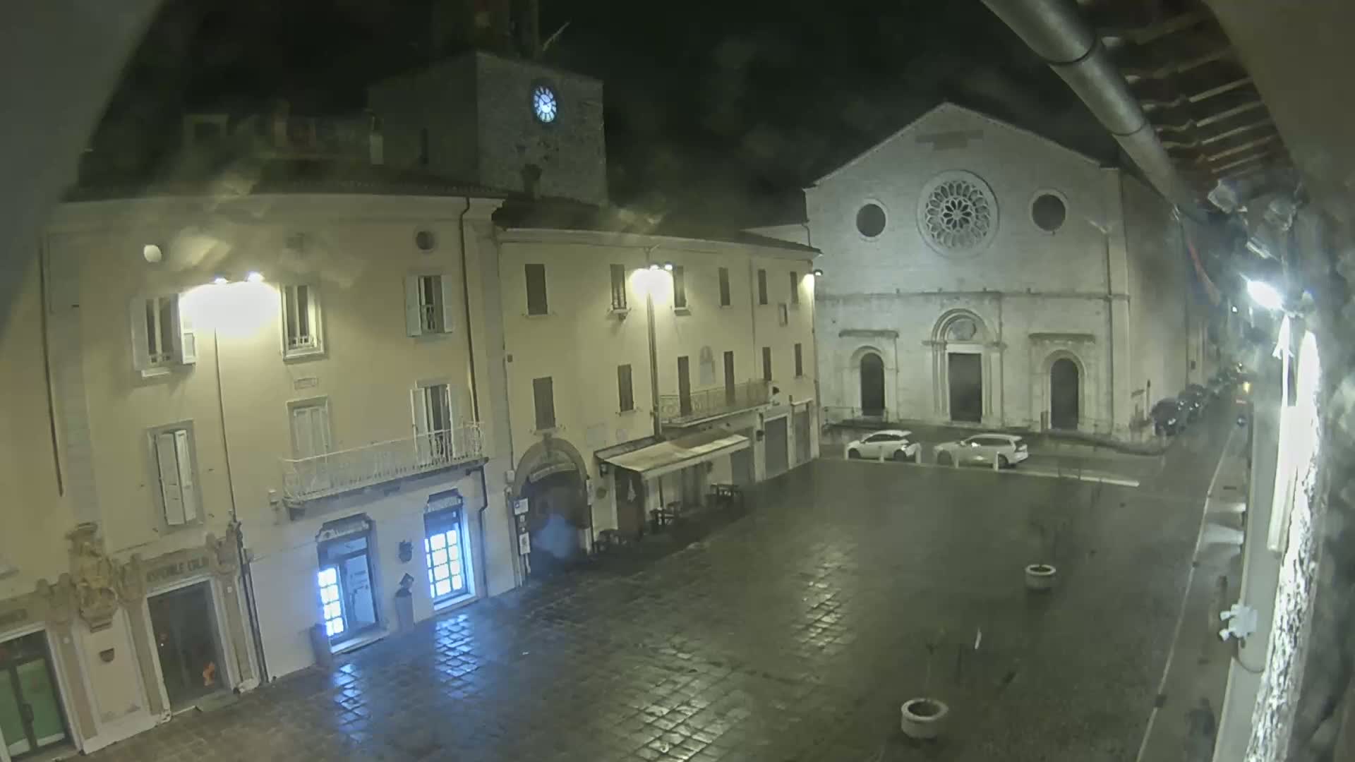 A nighttime view of a town square with several buildings, including a church with a rose window, under wet conditions.