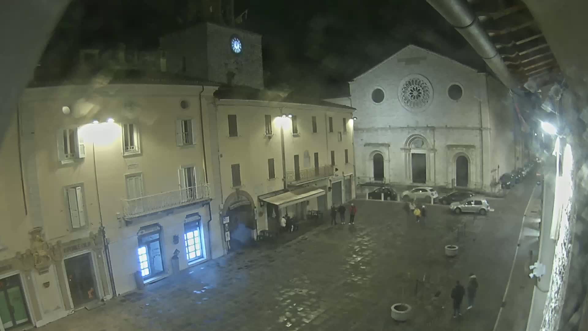 A nighttime view of a town square, featuring several buildings, a few parked cars, and several pedestrians under a cloudy sky.