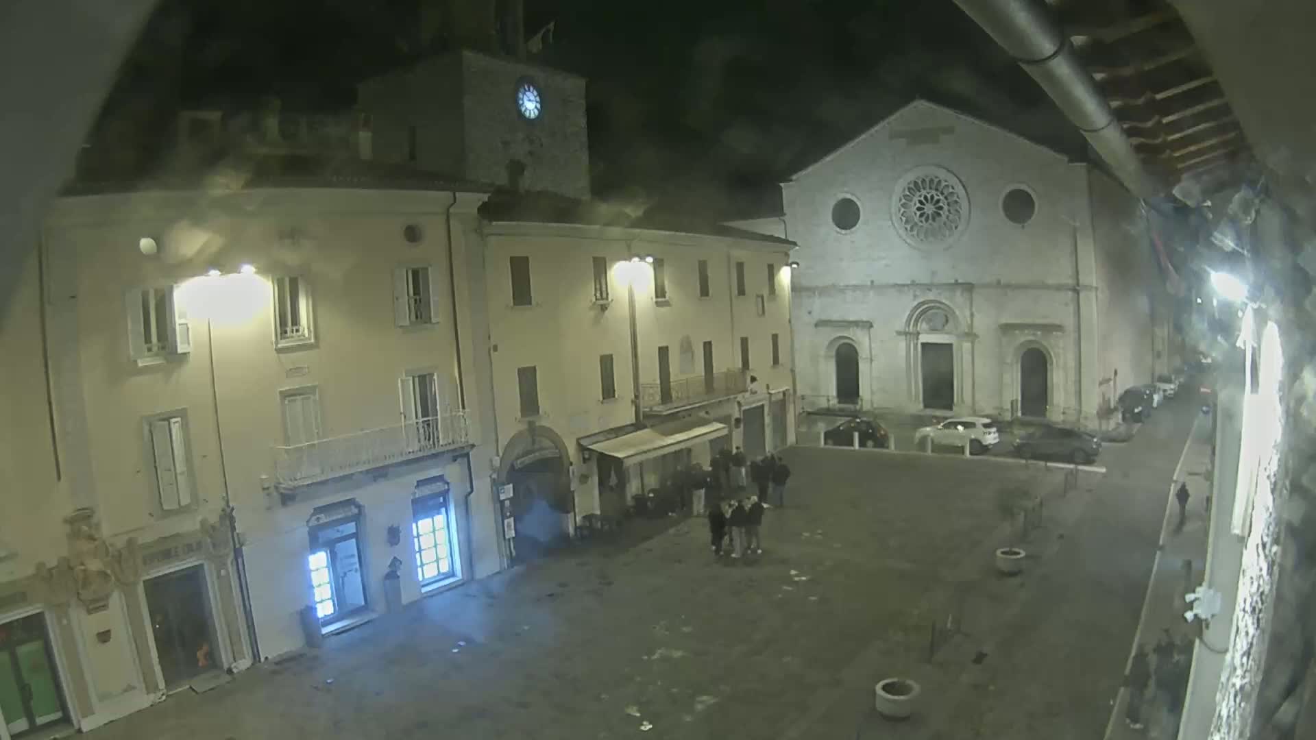 A nighttime, dimly lit town square features several buildings, including a church with a rose window, a clock tower, and a few shops, with a small group of people gathered near one of them.