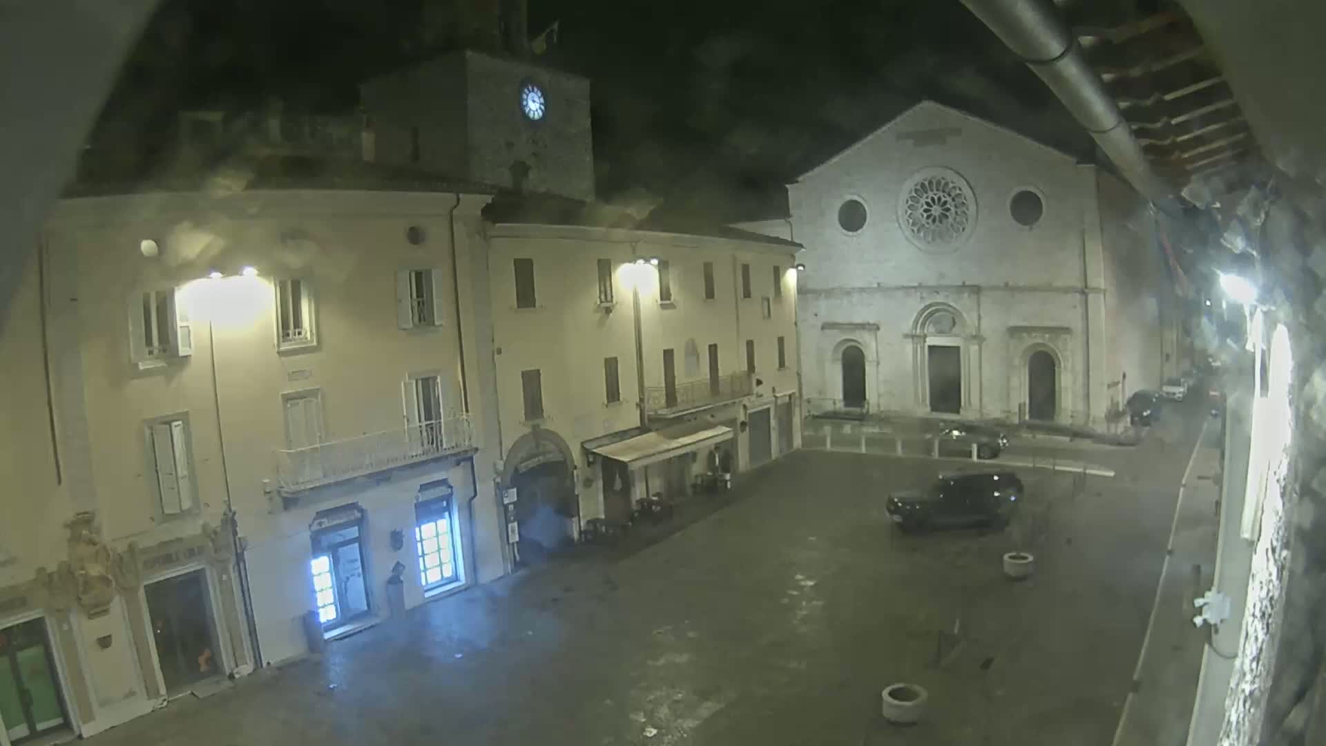 A nighttime, wet view of a town square with buildings, including a church with a rose window, and a few parked cars.
