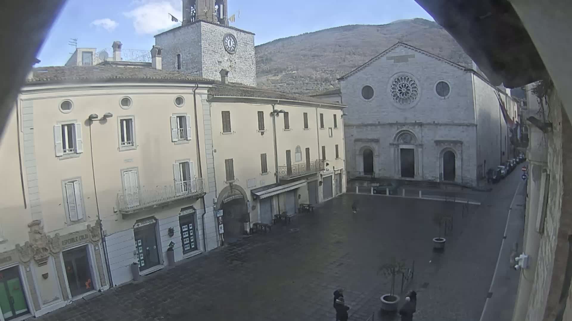 A mostly empty town square is seen on a cloudy day, with several buildings including a church and a clock tower visible, and a few people walking near potted plants.