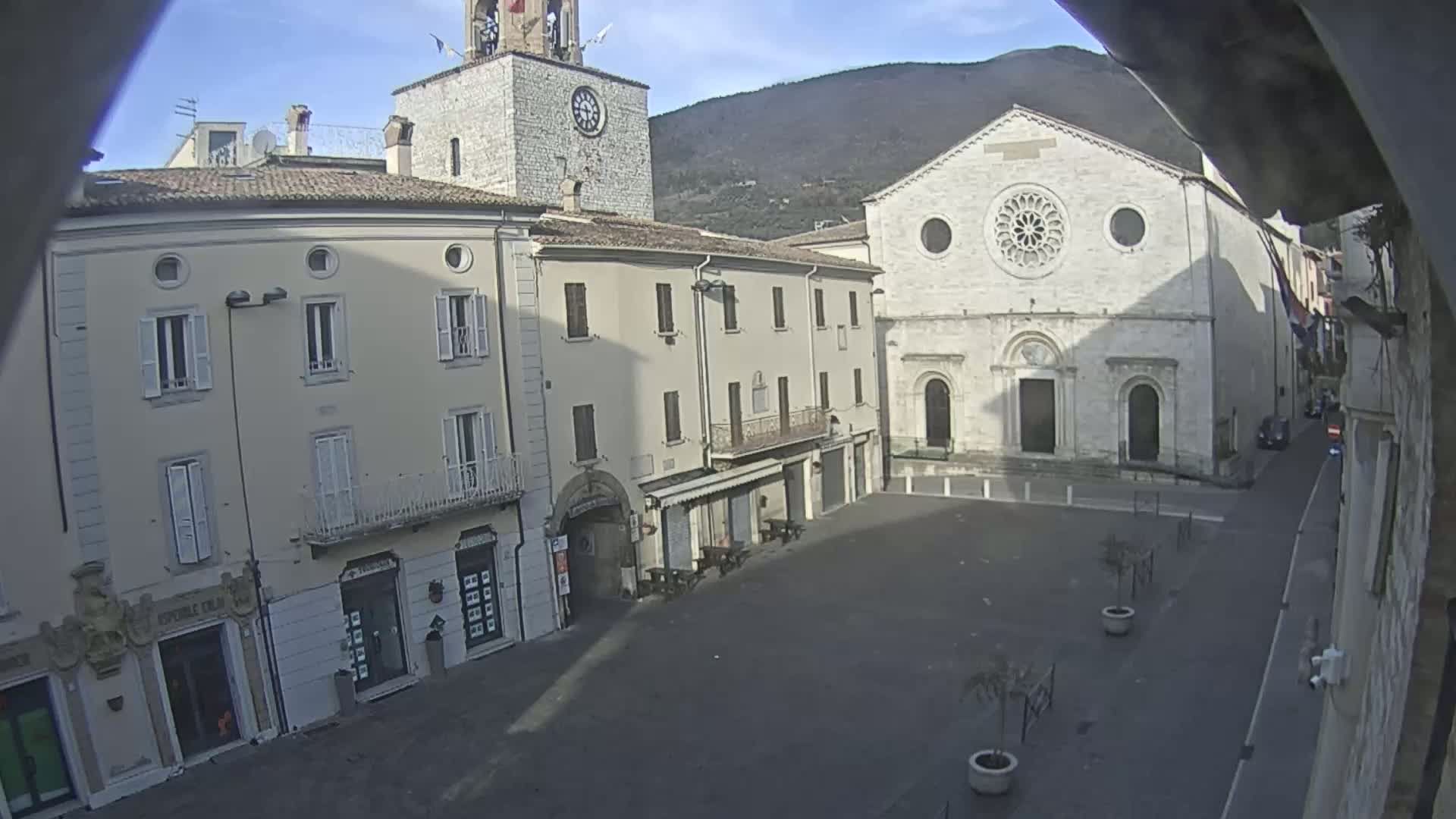 A mostly empty town square is viewed from above on a sunny day, featuring several light-colored buildings, including a church with a rose window and a clock tower.