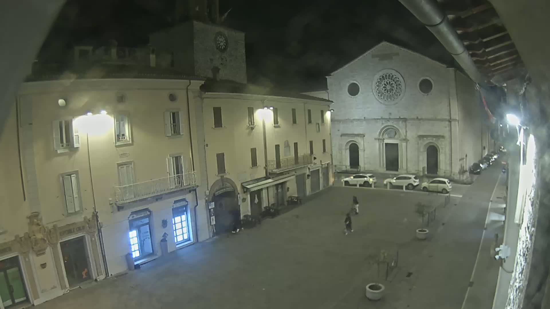 A nighttime view of a town square with several buildings, including a church with a rose window, under cloudy skies.