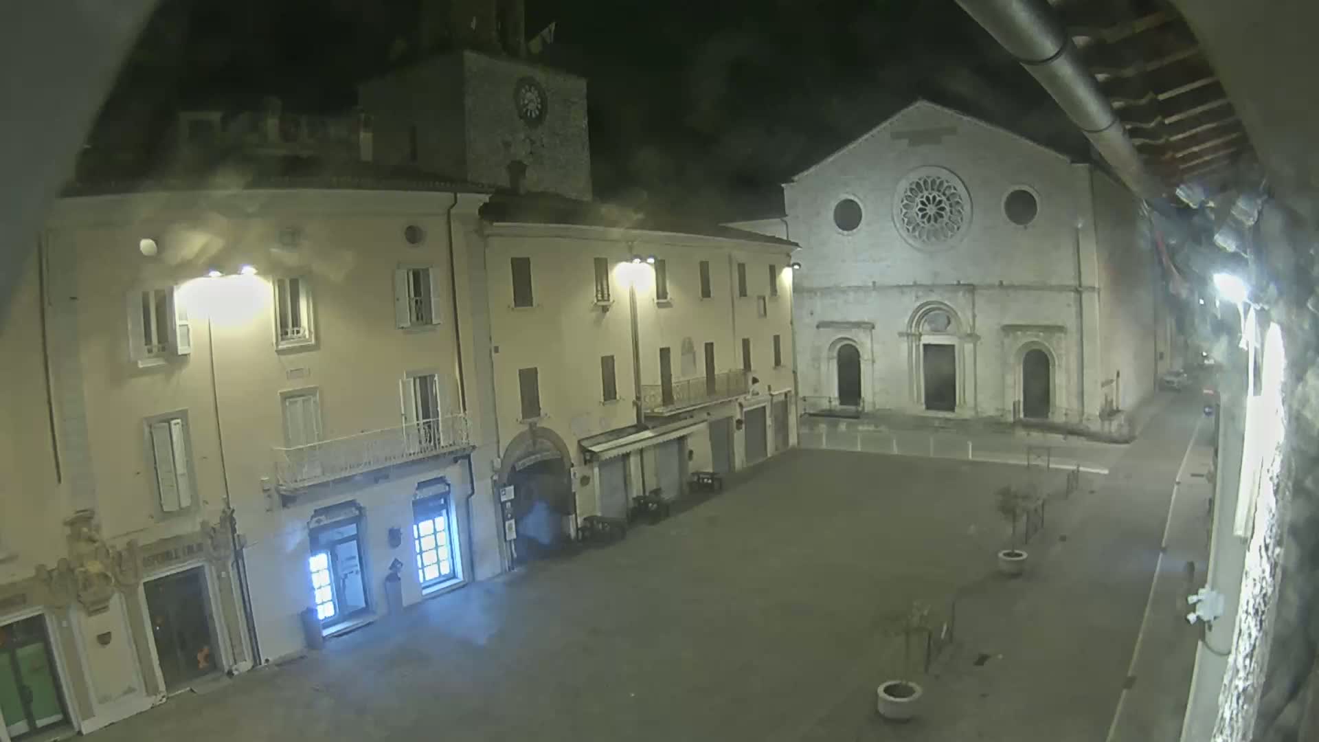 A nighttime view of a town square, showing several buildings including a church with a rose window, under slightly overcast conditions.