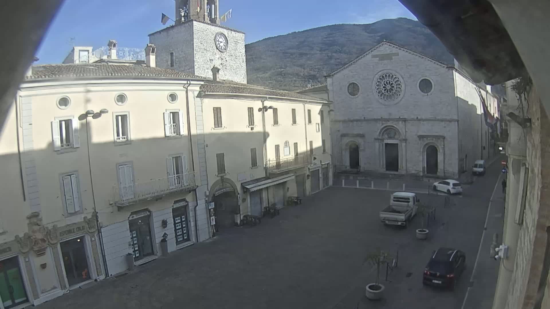 A sunny view of a town square with several buildings, including a church and a clock tower, a few cars parked, and some small trees in planters.