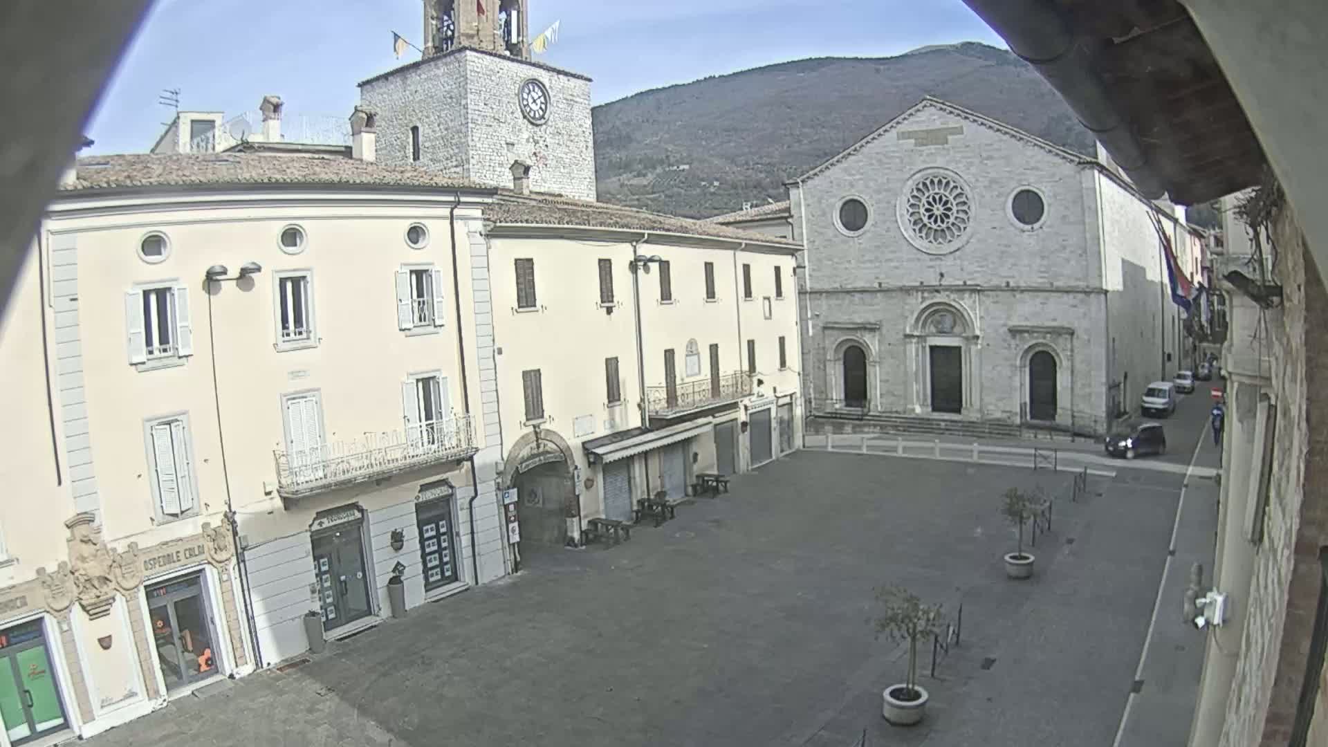 A mostly empty town square is viewed from above on a clear day, showing several buildings including a church with a rose window and a clock tower, with mountains visible in the background.