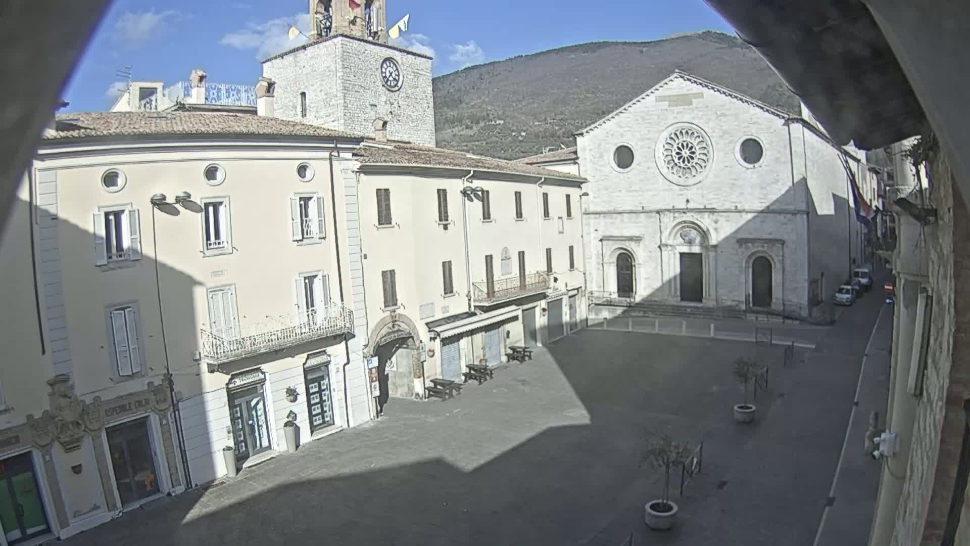 A sunny day reveals a town square with several buildings, including a clock tower and a large church with a rose window, backed by mountains.