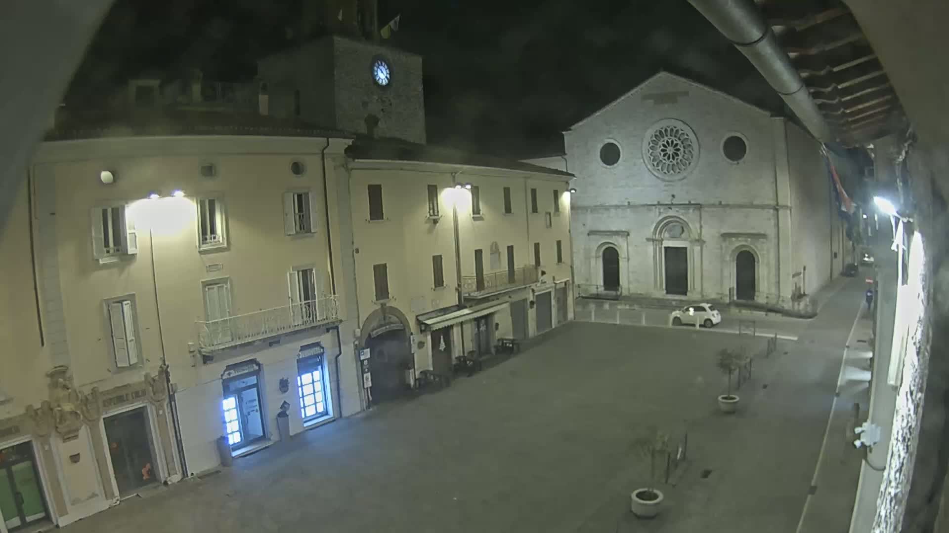 A nighttime view of a town square, featuring a clock tower, a large stone church, and several buildings, under a dark sky.
