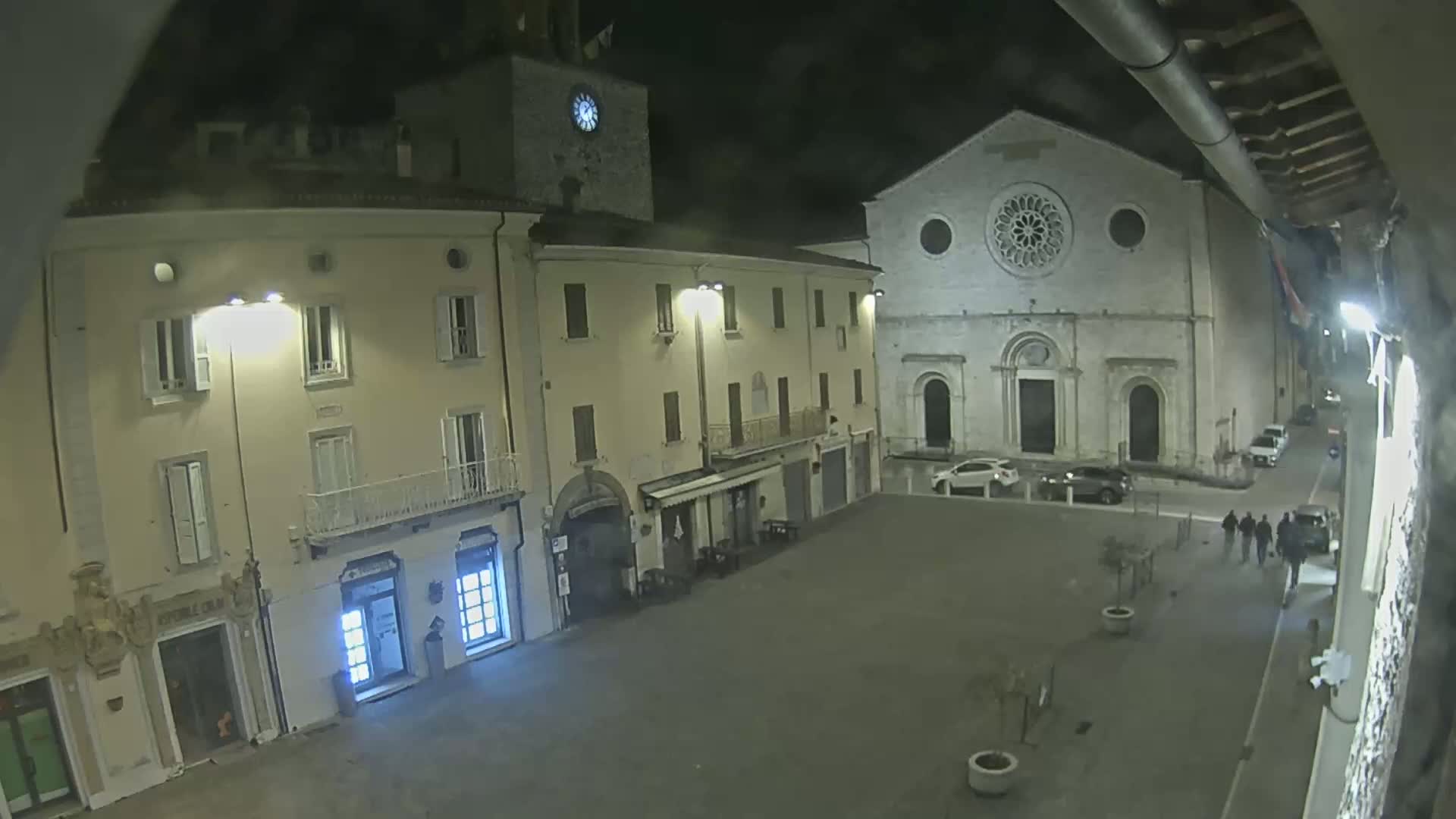 A nighttime view of a town square, showing several buildings, including a church with a rose window, under overcast skies.