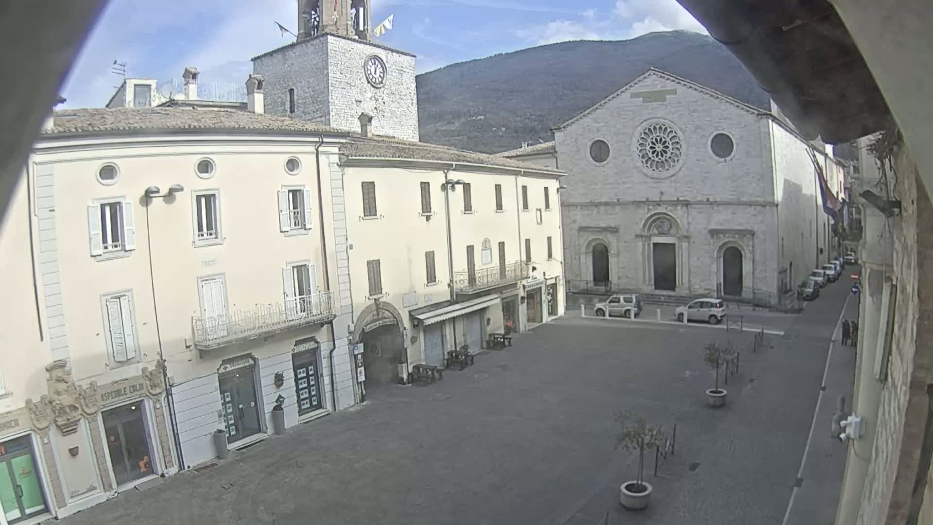 A mostly empty town square is seen on a sunny day, with light-colored buildings surrounding it, including a church with a large rose window and a clock tower.