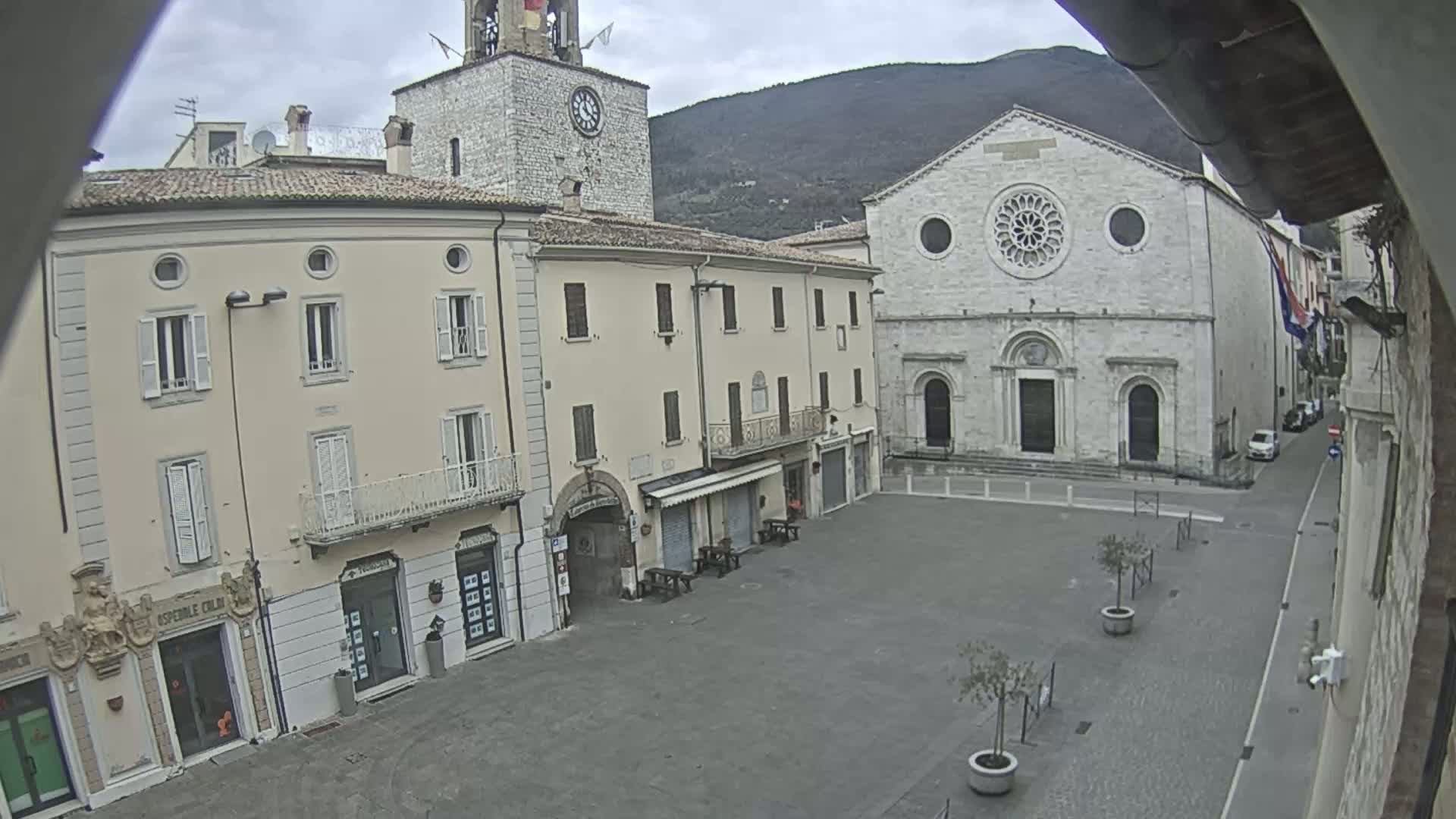 A mostly empty town square is surrounded by light-colored buildings, including a church with a rose window, under an overcast sky.