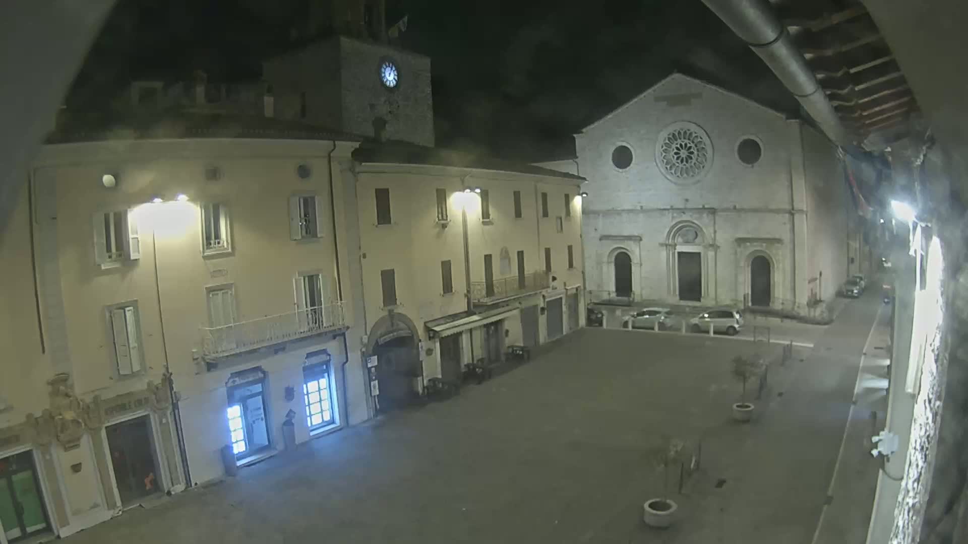 A nighttime view of a town square, featuring several buildings including a church with a large rose window, under a partly cloudy sky.