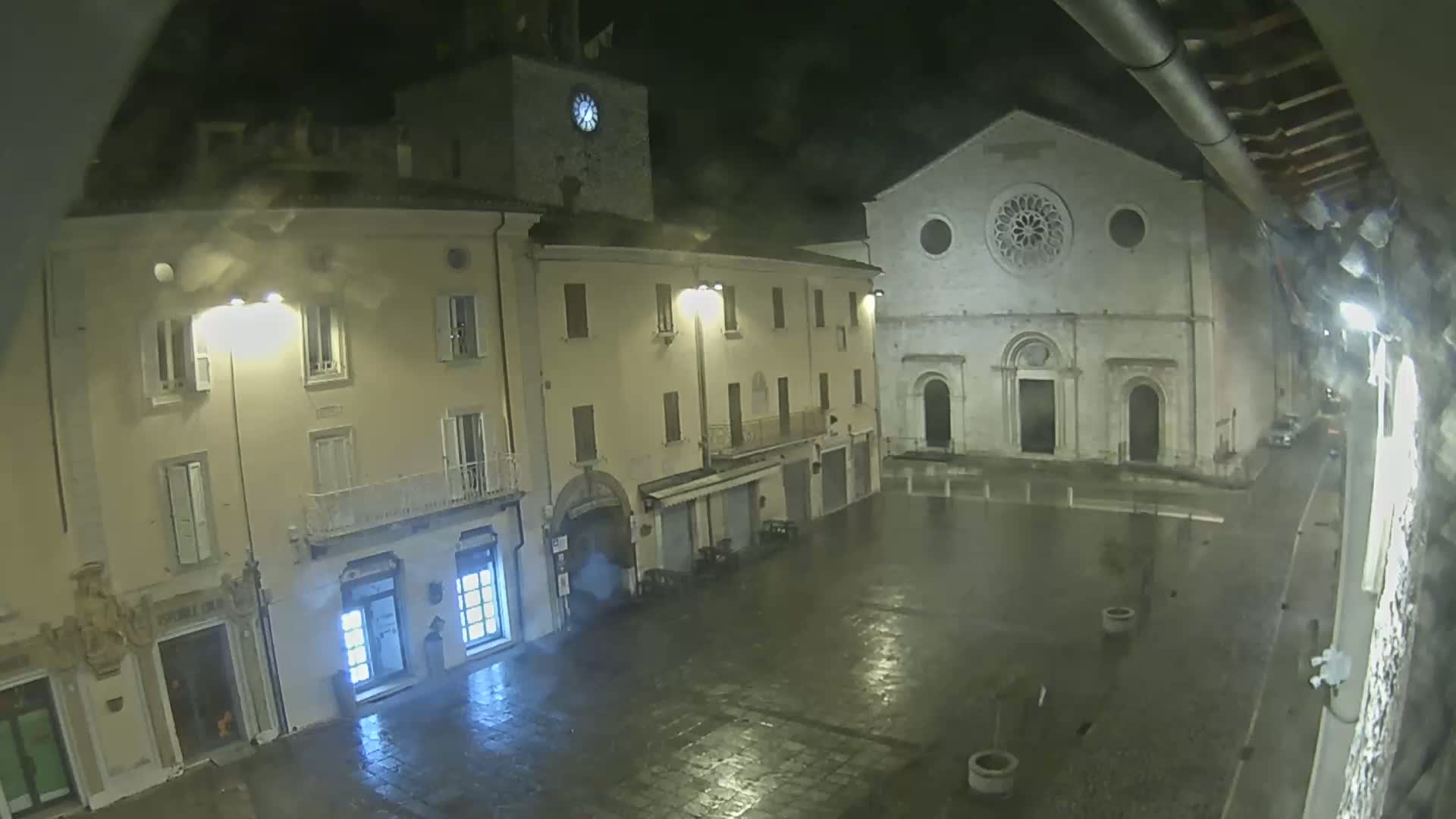A nighttime view of a rain-soaked town square, showing illuminated buildings including a church with a rose window and a clock tower.