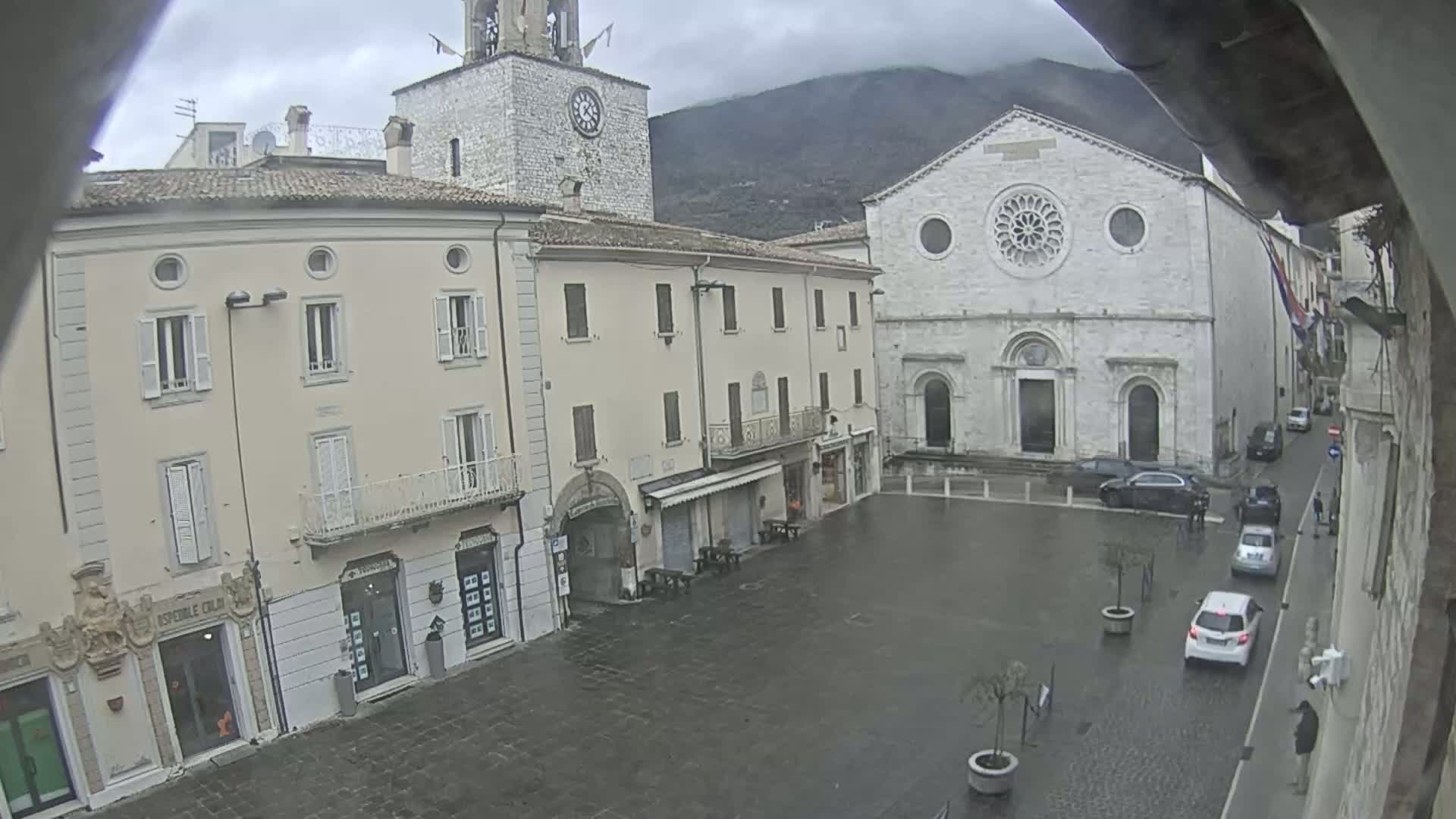 A town square with several buildings, including a church with a rose window, is seen on an overcast day, with cars parked and a few pedestrians present.