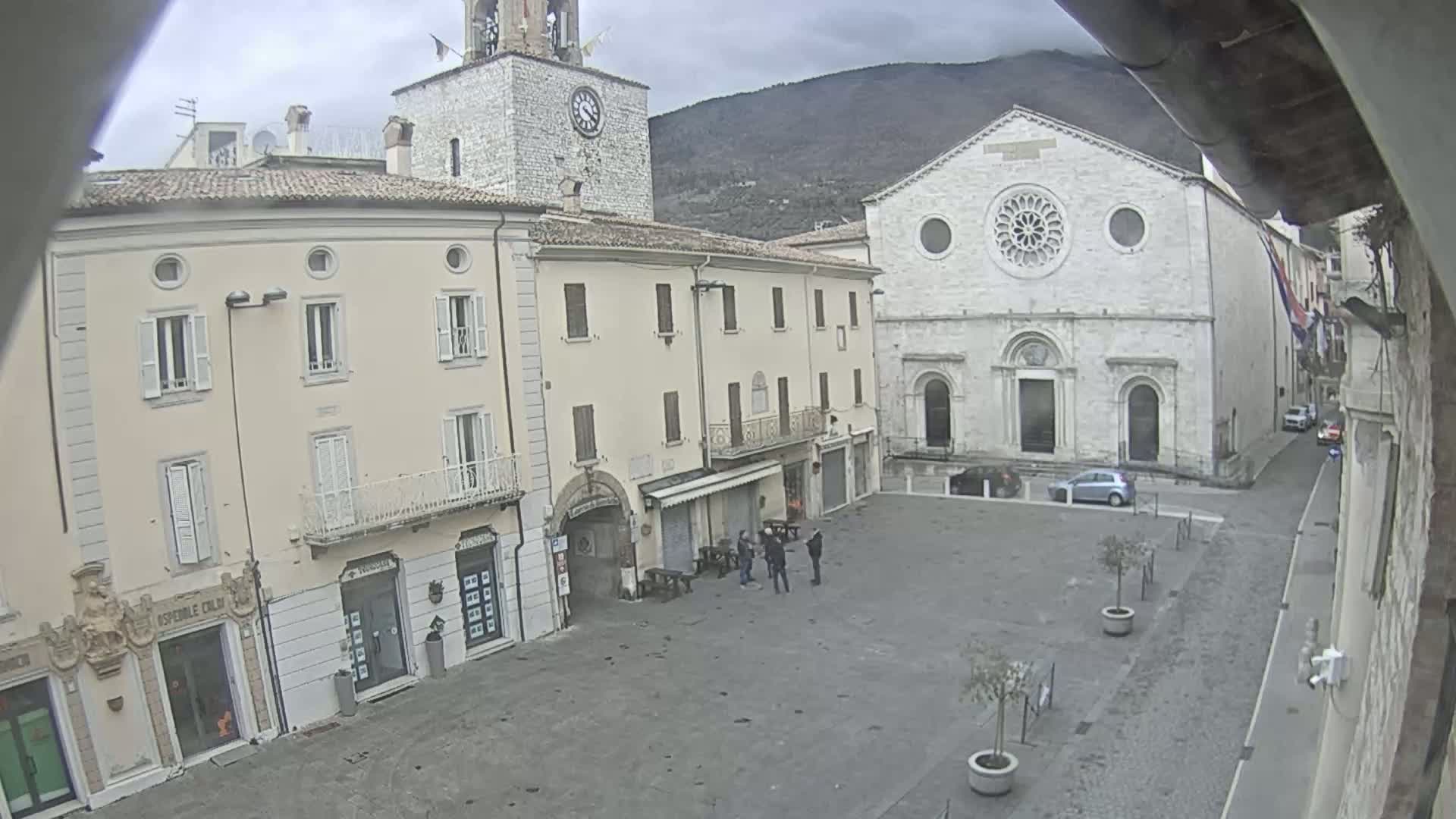 A town square with several buildings, including a church with a rose window and a clock tower, under an overcast sky.